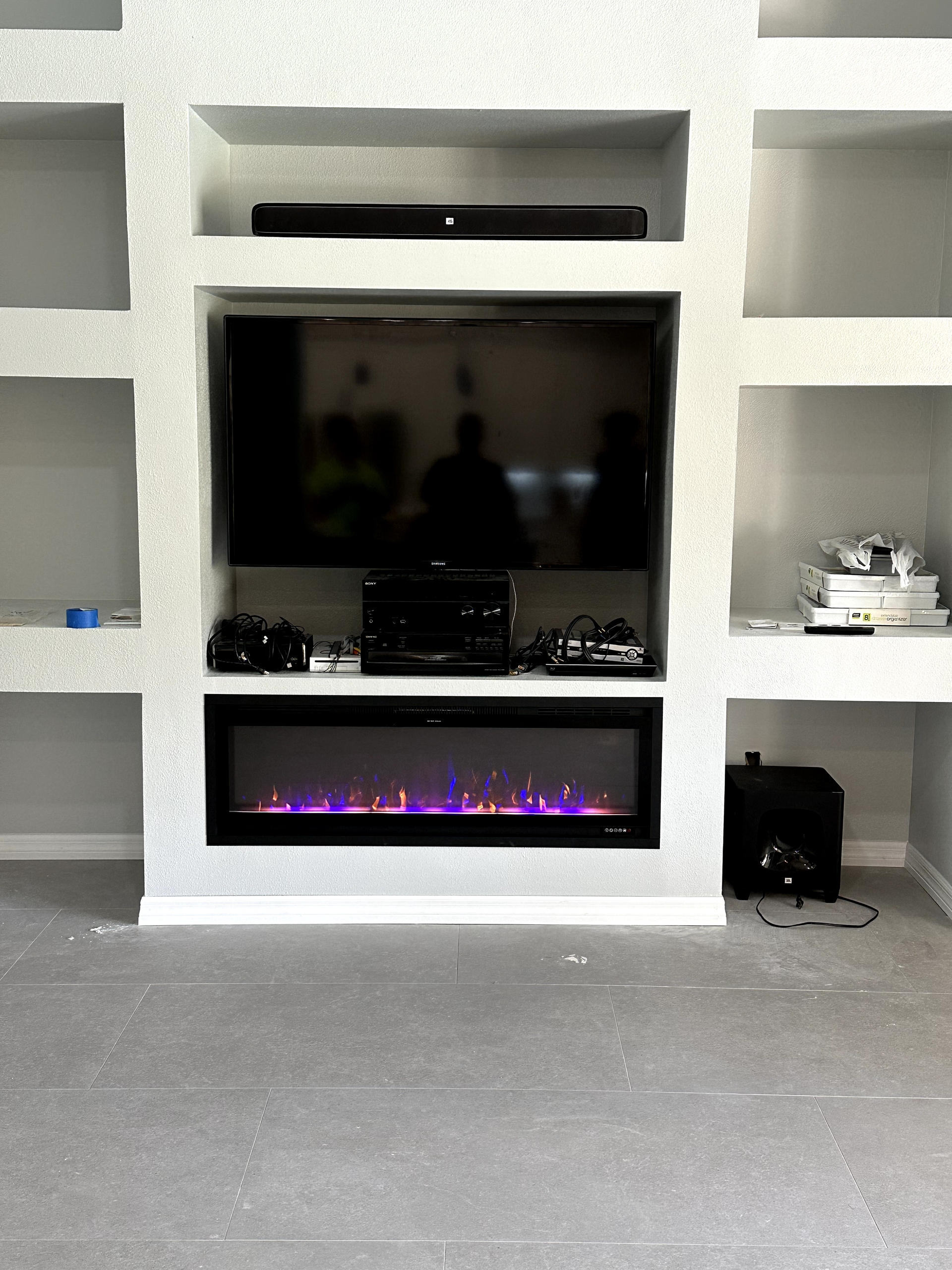 Built-in entertainment center with fireplace, TV, soundbar, and shelves. Displaying a lit fireplace with purple flames.