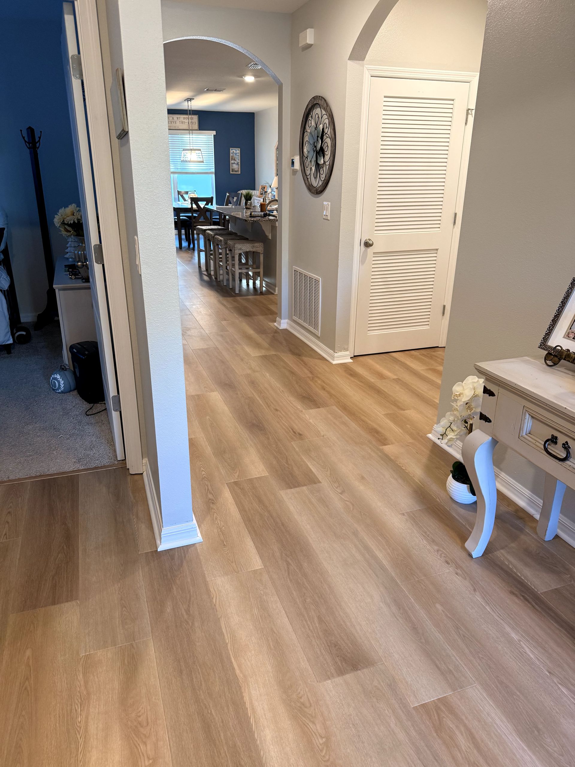 Hallway with light wood-look flooring, arched doorway leading to kitchen, white door with shutter design, and small table.
