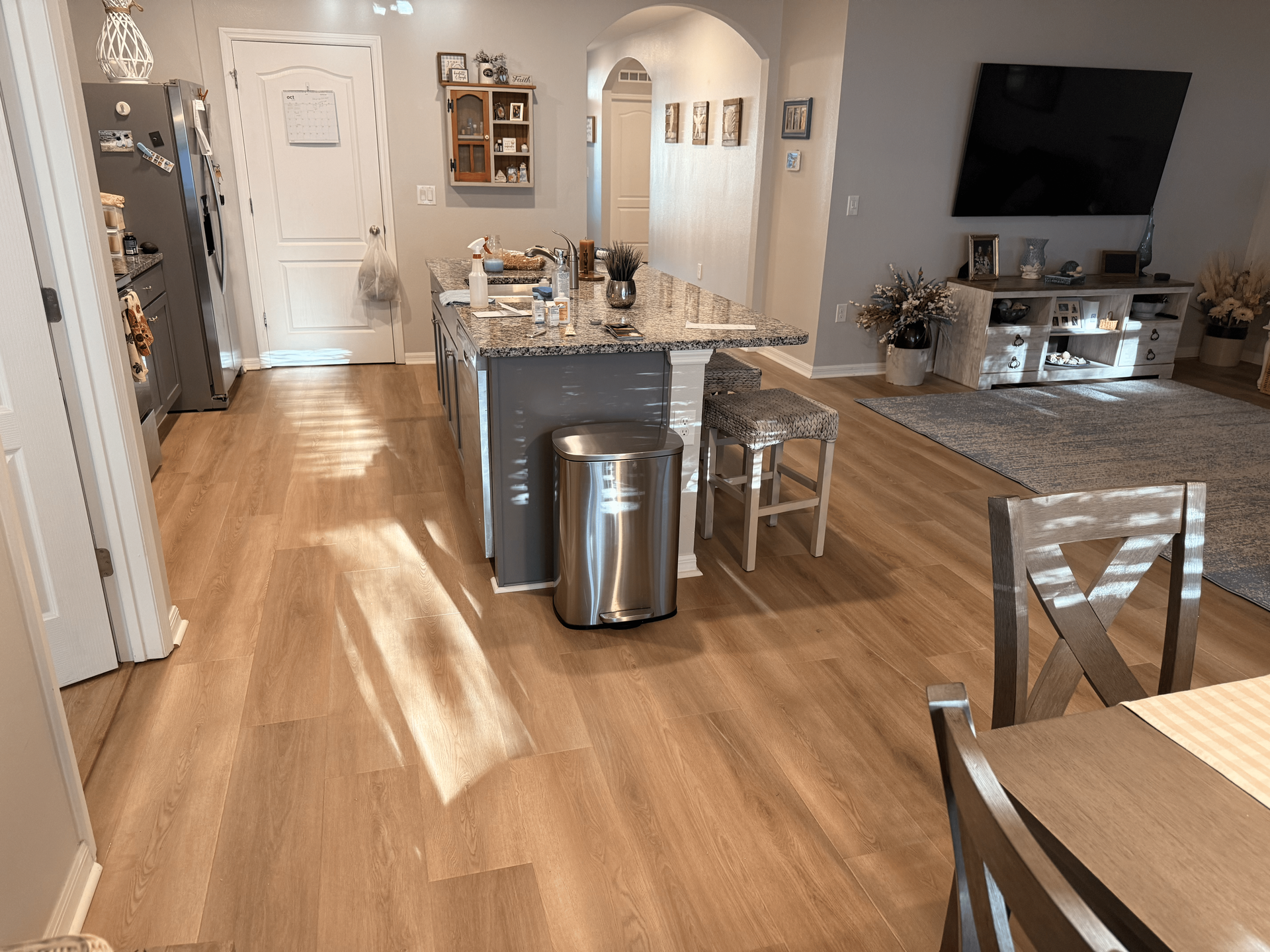 Kitchen with wood-look flooring, island with stools, stainless steel trash can, and TV in the background.
