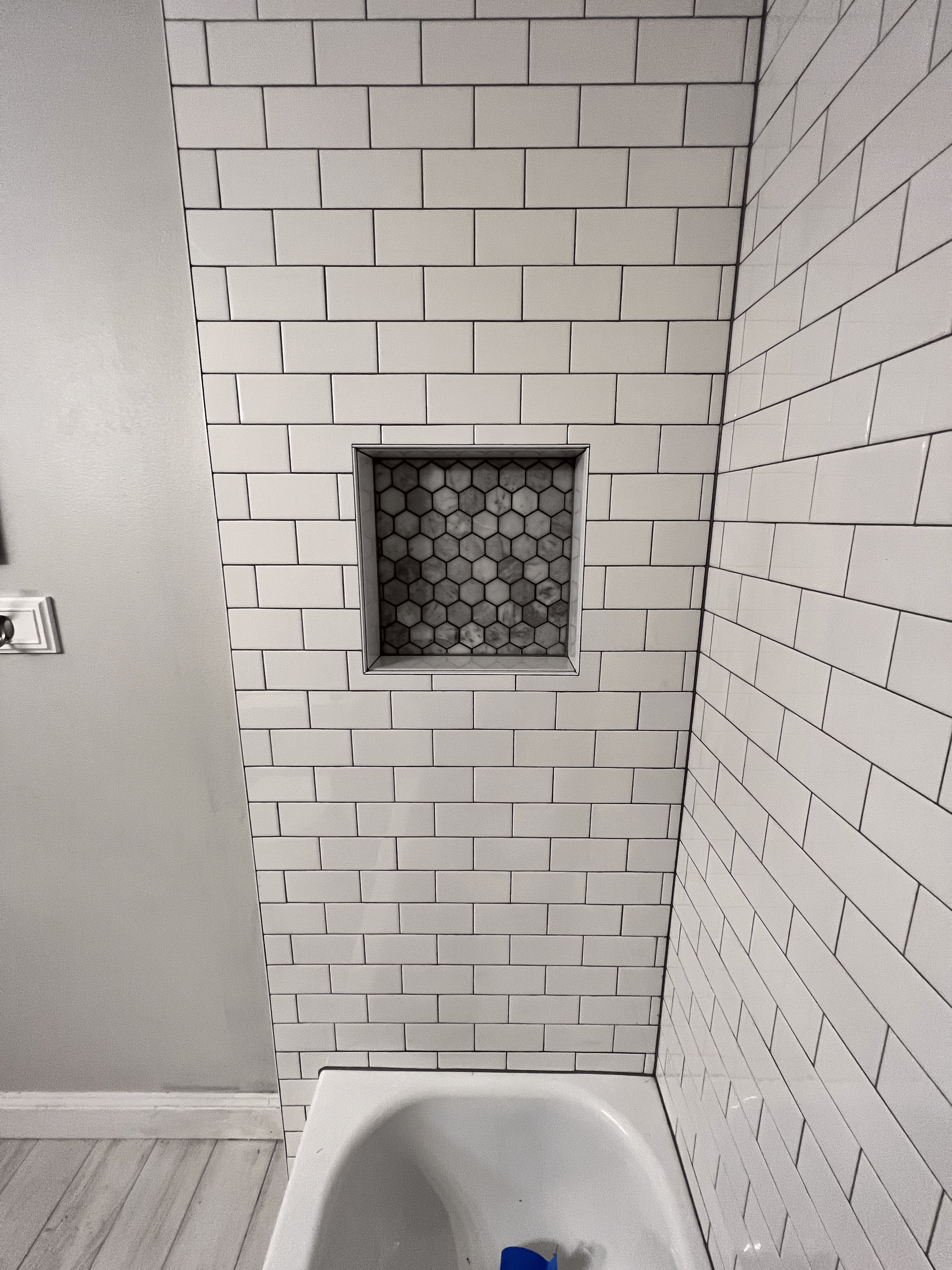 White tiled shower wall with a built-in recessed shelf filled with gray hexagon tiles, above a bathtub.