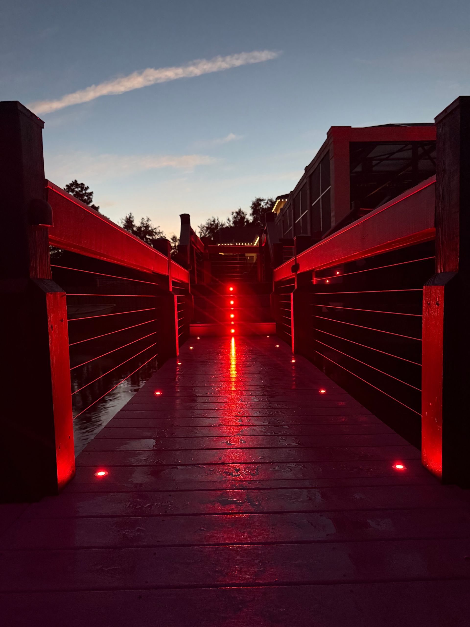 Red-lit bridge over water at dusk. Wooden walkway with red lights on both sides, leading toward a distant structure.