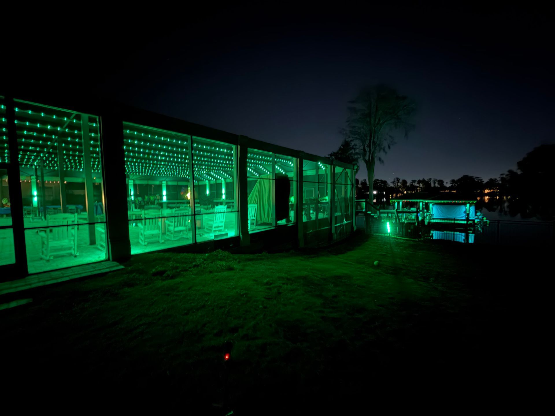 Green-lit building at night, next to water. Exterior lighting reflects off the grass and structures, with a dark sky.