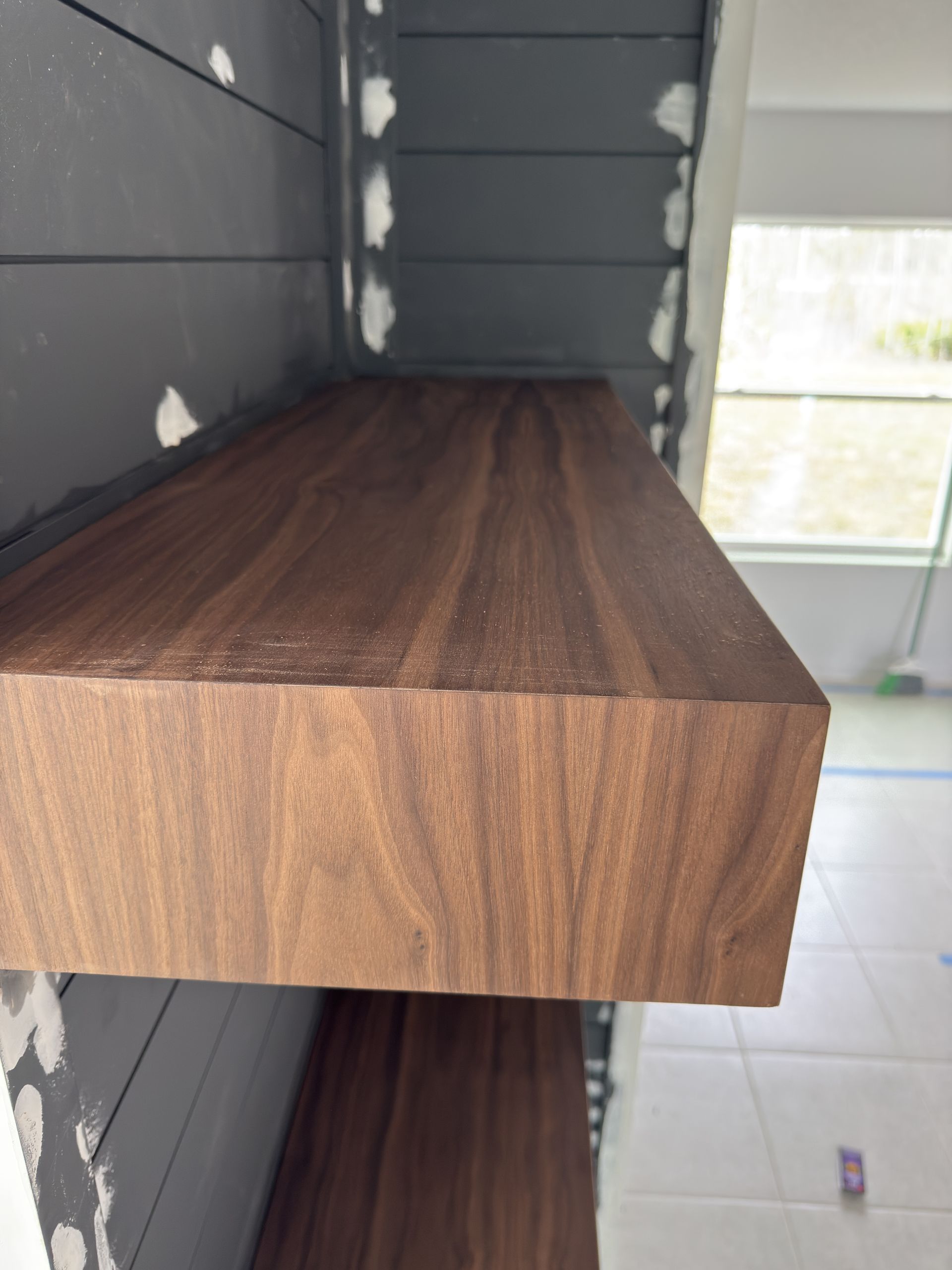 A brown wooden shelf attached to a gray wall with visible wood grain, near a window.