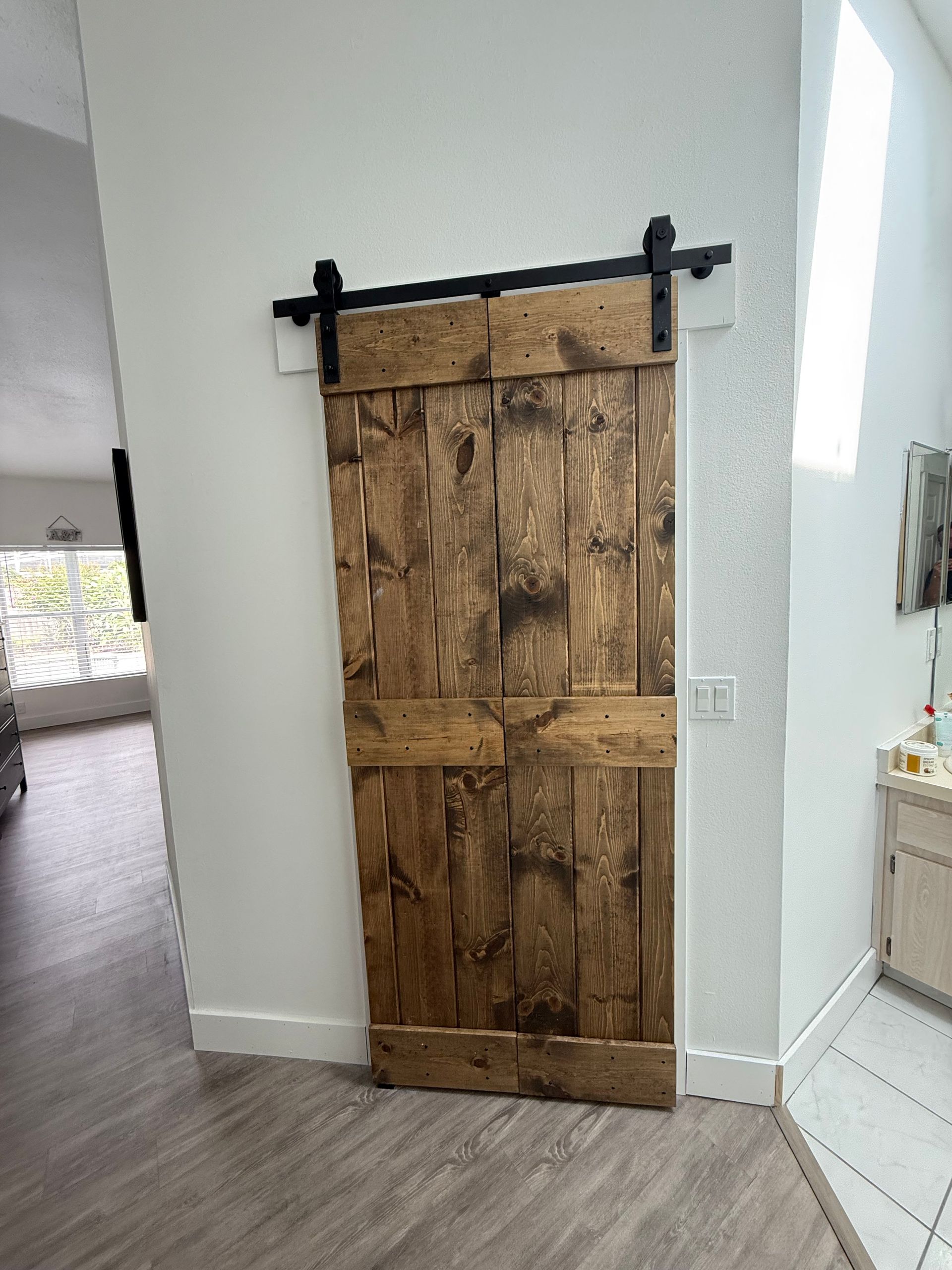 Barn door, mounted on a black track, is made of stained wood, against a white wall.