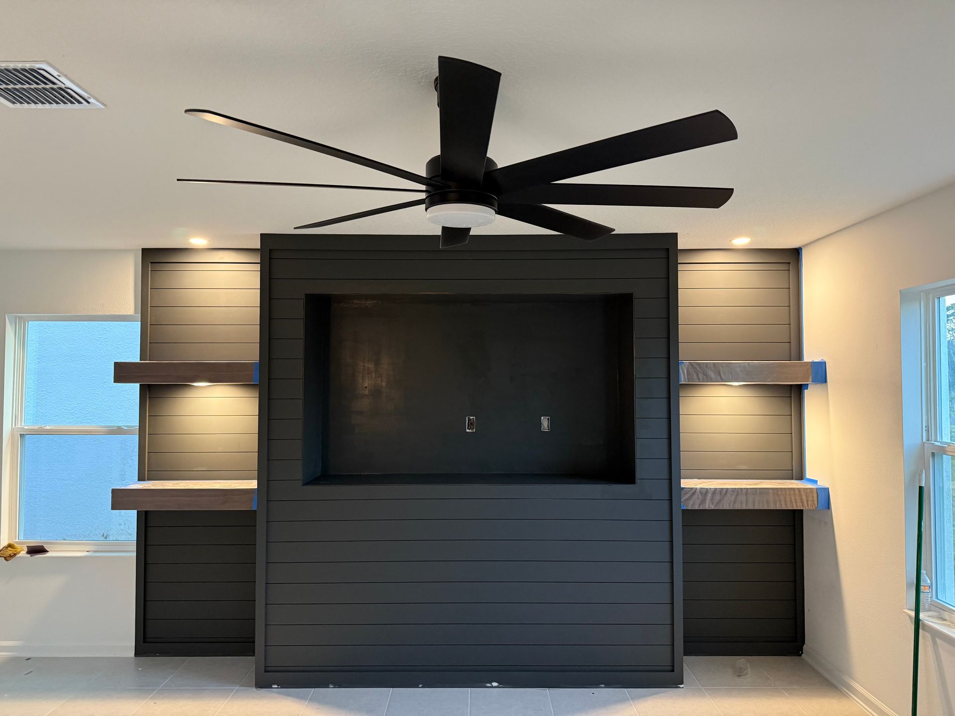 Dark gray media center with TV space, shelves, and a black ceiling fan in a room.