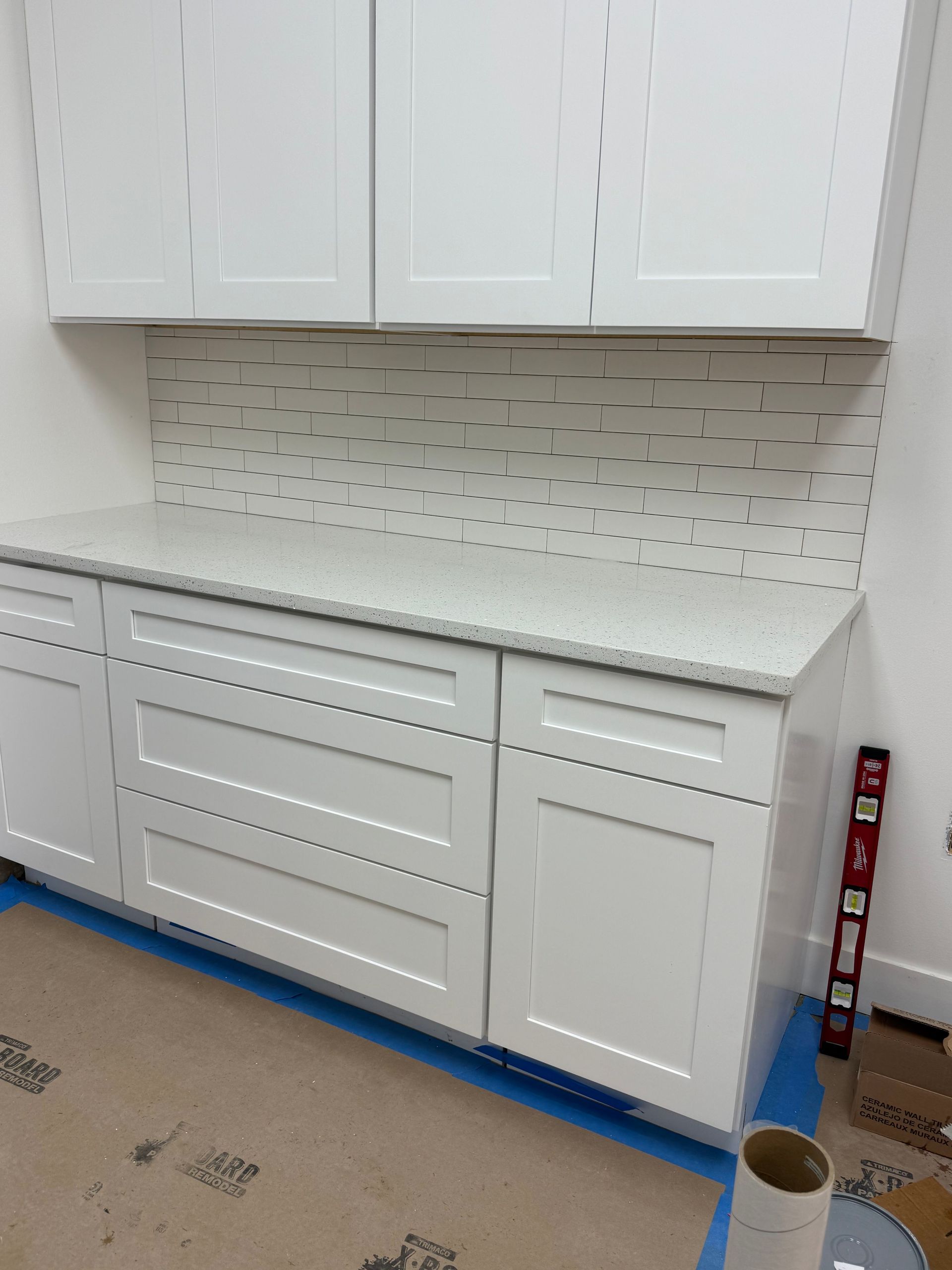 White kitchen cabinets, countertop, and tiled backsplash installed. A level leans against the wall.