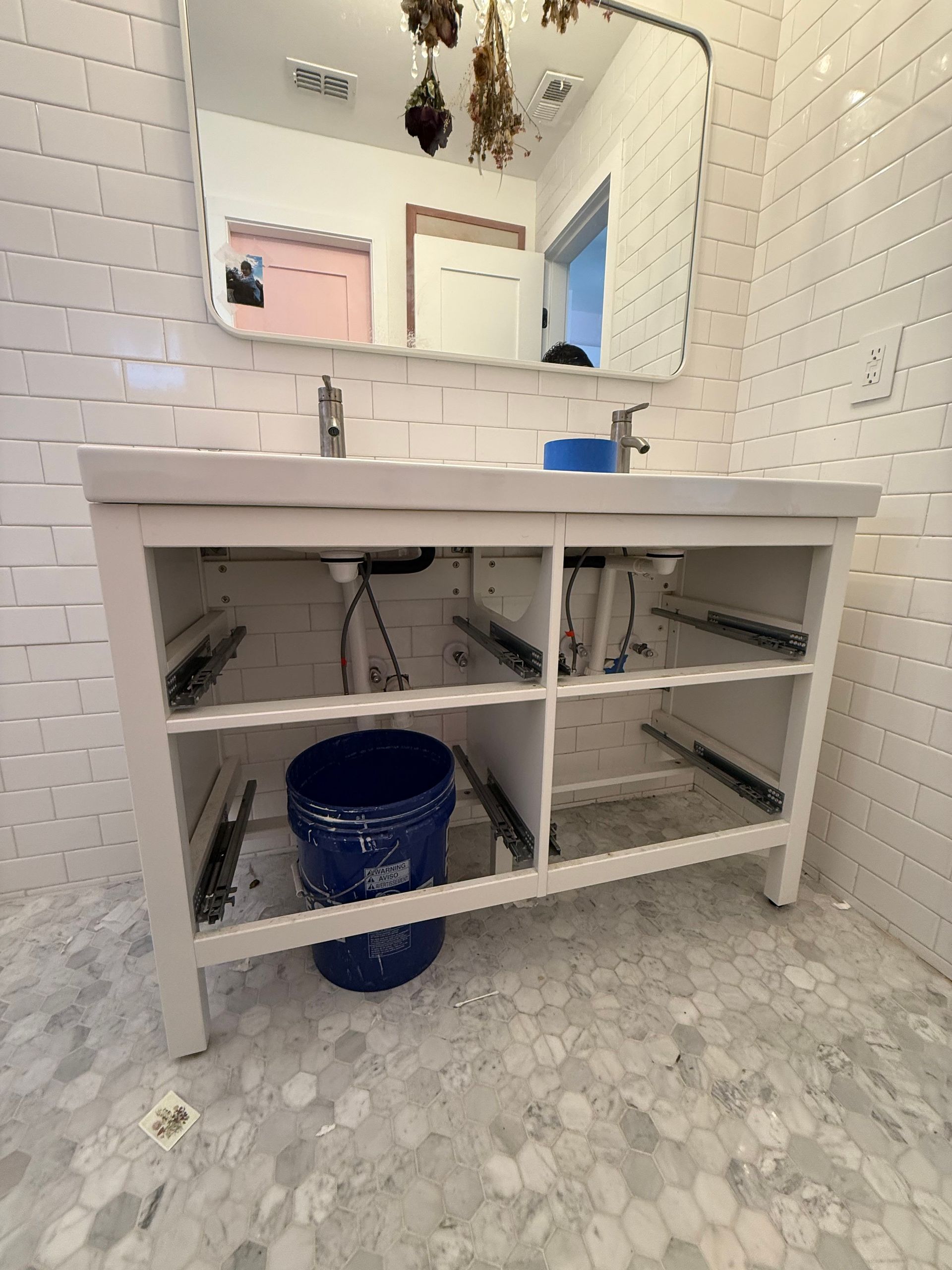 Bathroom vanity with open storage, mirror, white subway tile, and a blue bucket.