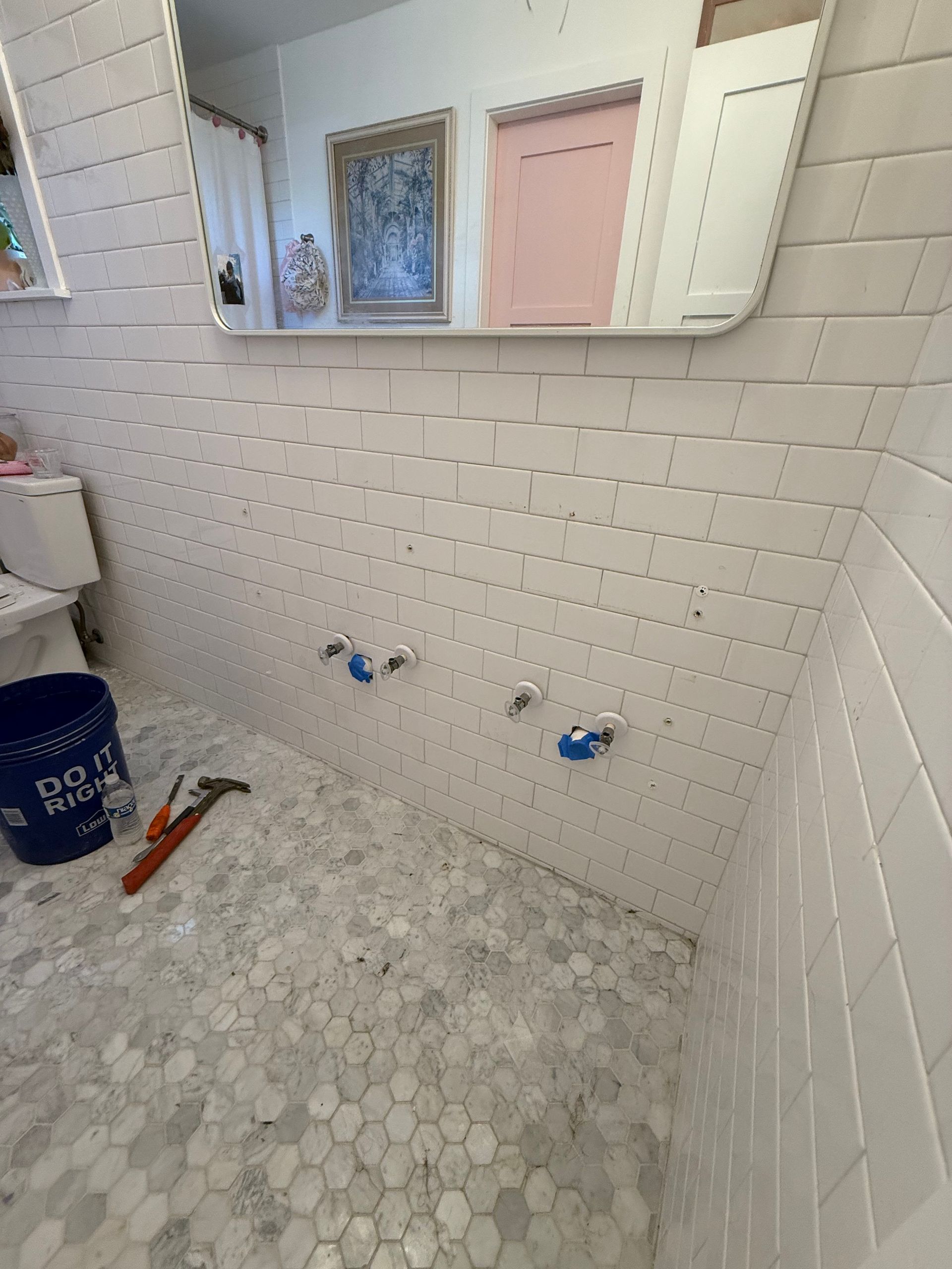 Bathroom under construction with white tiled walls and floor, blue water valves, a mirror, and tools.