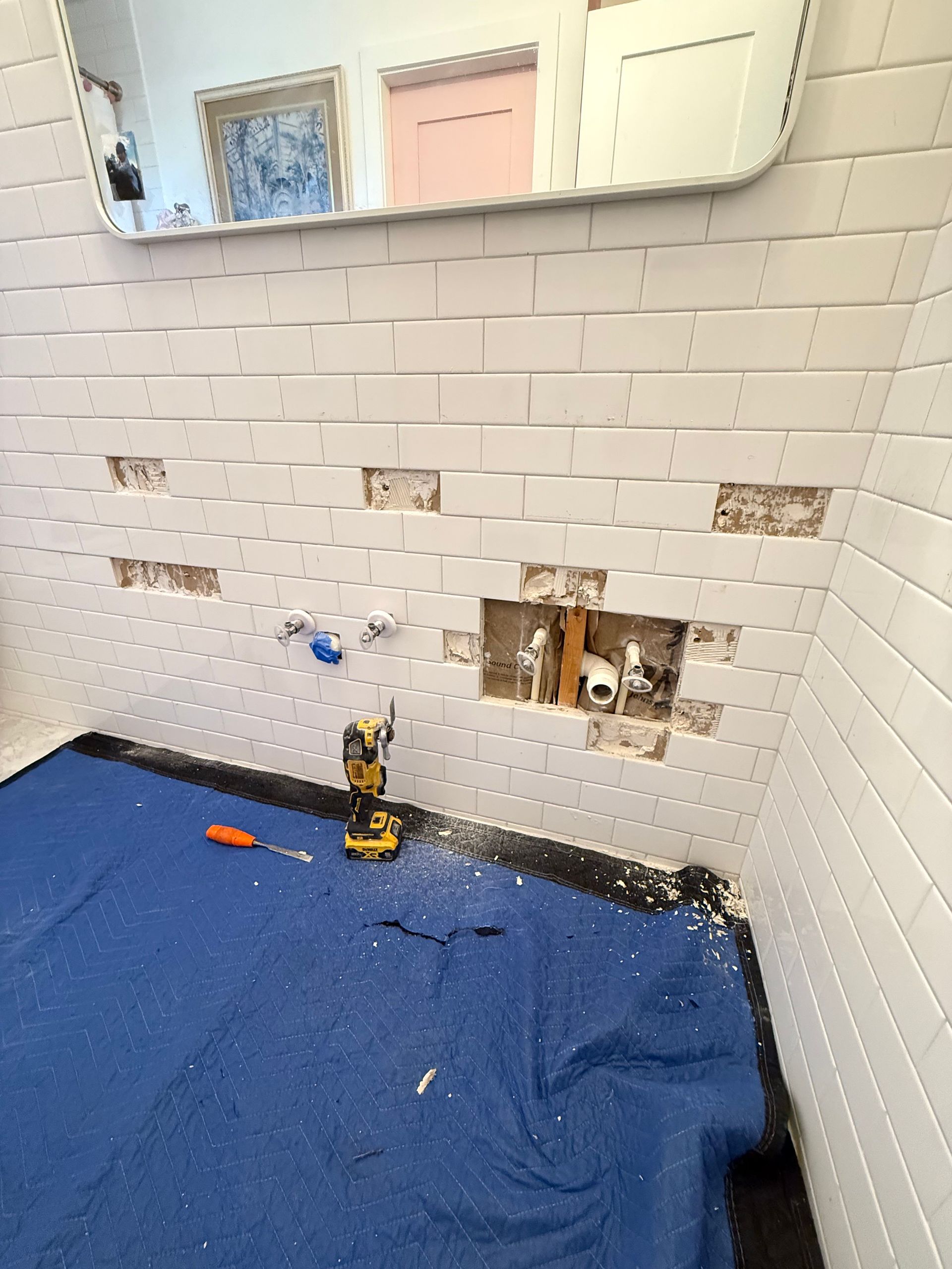 Bathroom wall with damaged tiles, exposed plumbing, a drill, and blue drop cloth.