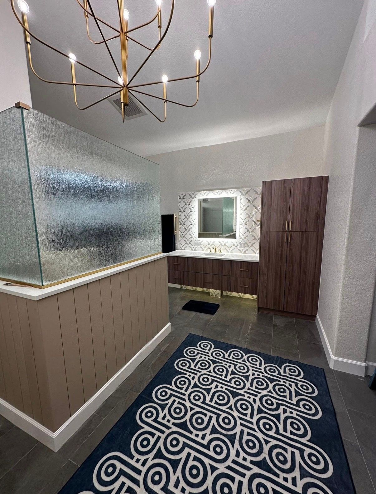 Modern bathroom with a patterned rug, large glass divider, and gold chandelier.