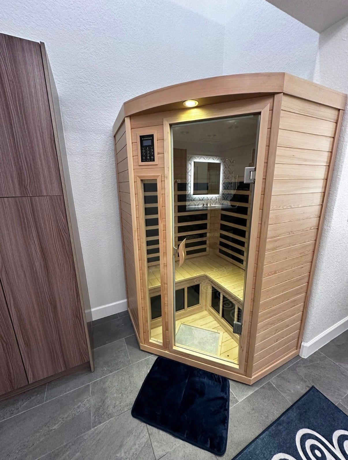 Wooden sauna in a room next to a brown wardrobe and a dark blue rug.
