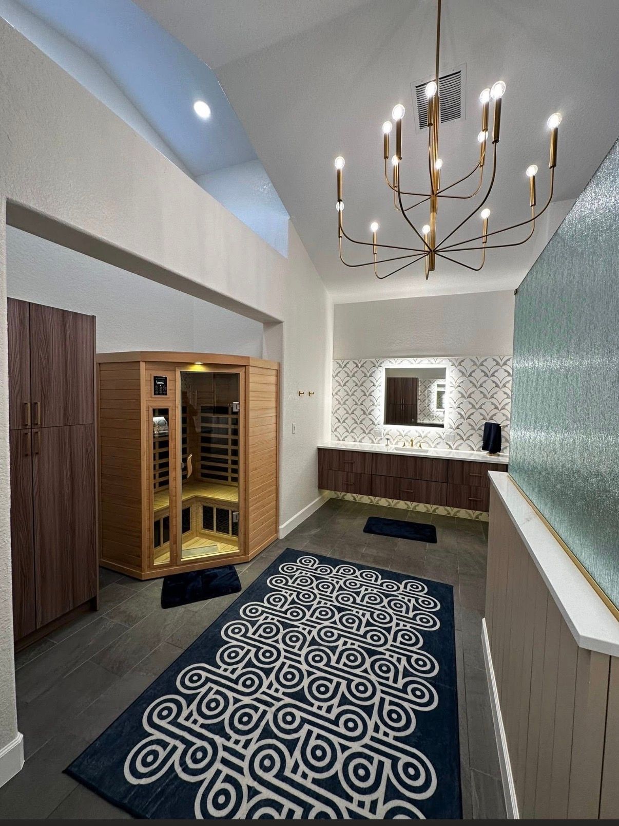 Modern spa-like bathroom with sauna, dark wood cabinets, blue patterned rug, and chandelier.