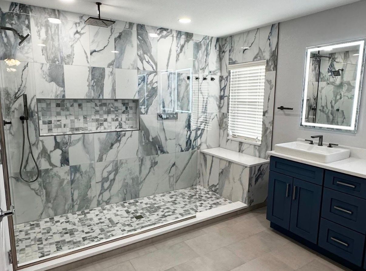 Modern bathroom with marble-look tile, a walk-in shower, a blue vanity, and a lighted mirror.