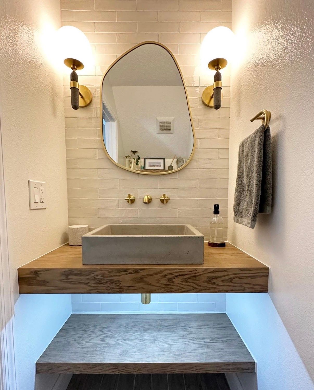 Modern powder room with floating wood vanity, concrete sink, gold fixtures, and irregular mirror.