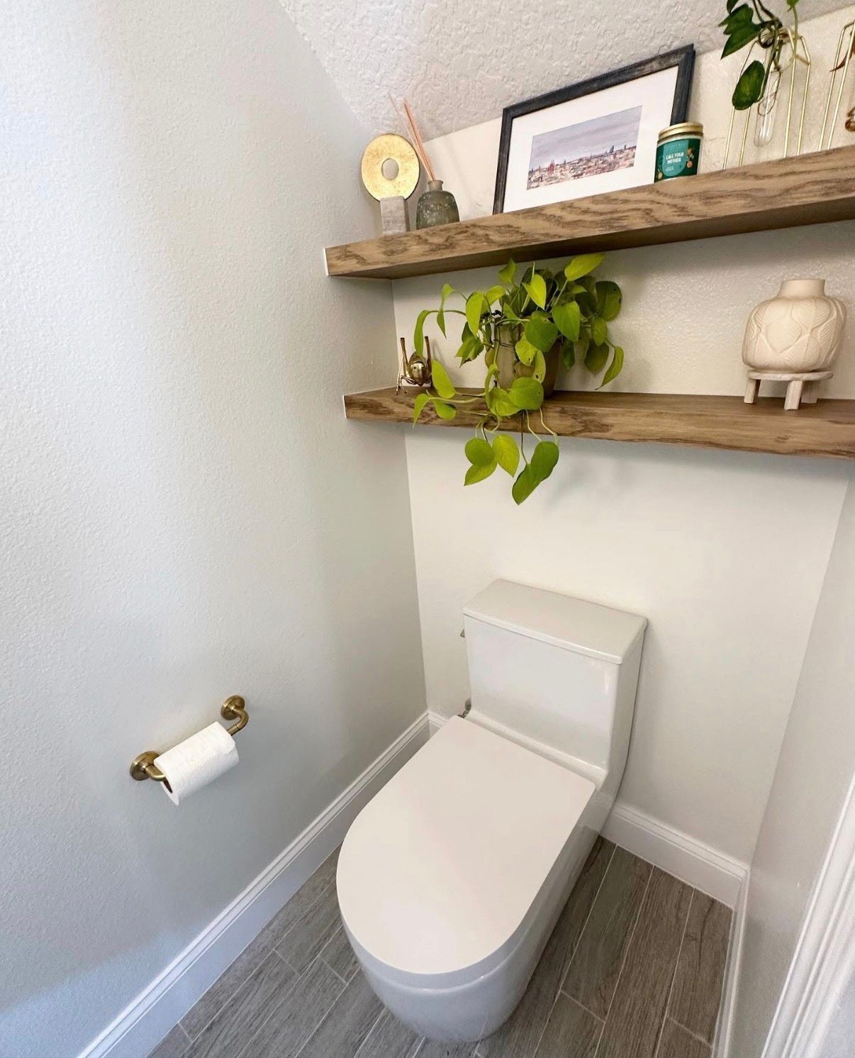 A small, modern bathroom with a toilet and wooden shelves. The shelves hold plants and decor.