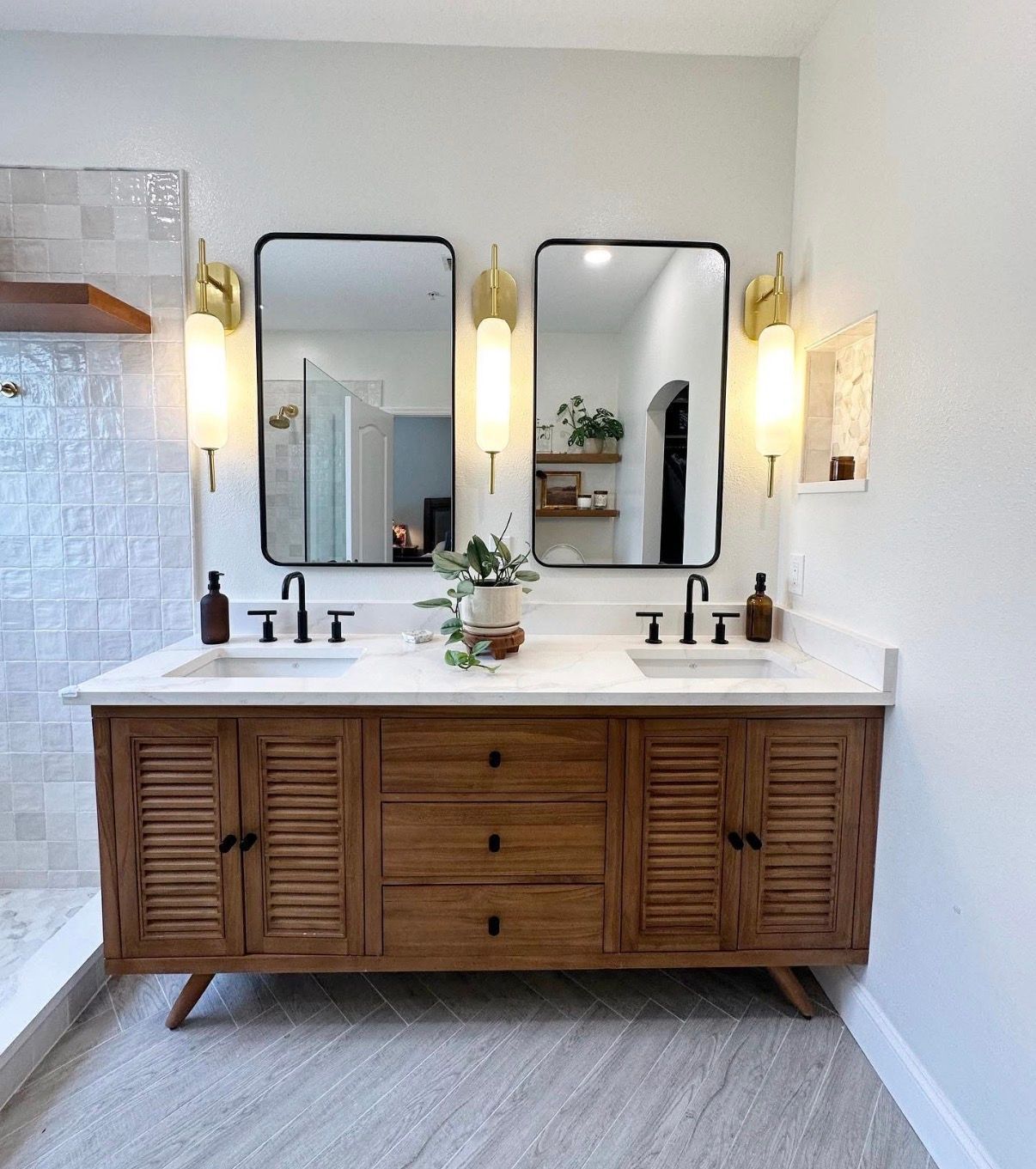 Bathroom vanity with two mirrors, gold sconces, wooden cabinet, black faucets, and white countertop.
