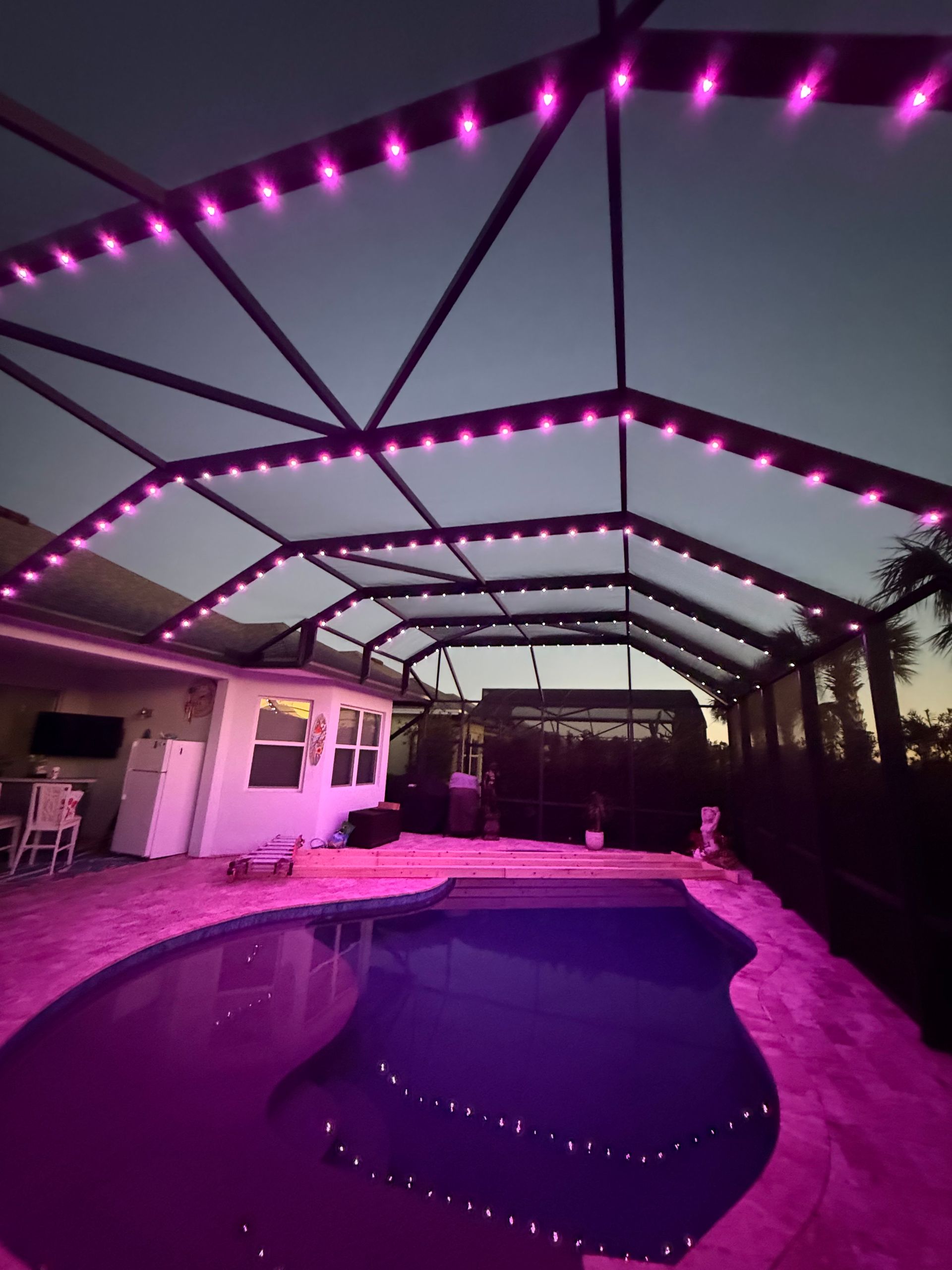 Swimming pool at dusk, lit with pink lights on a screened enclosure.
