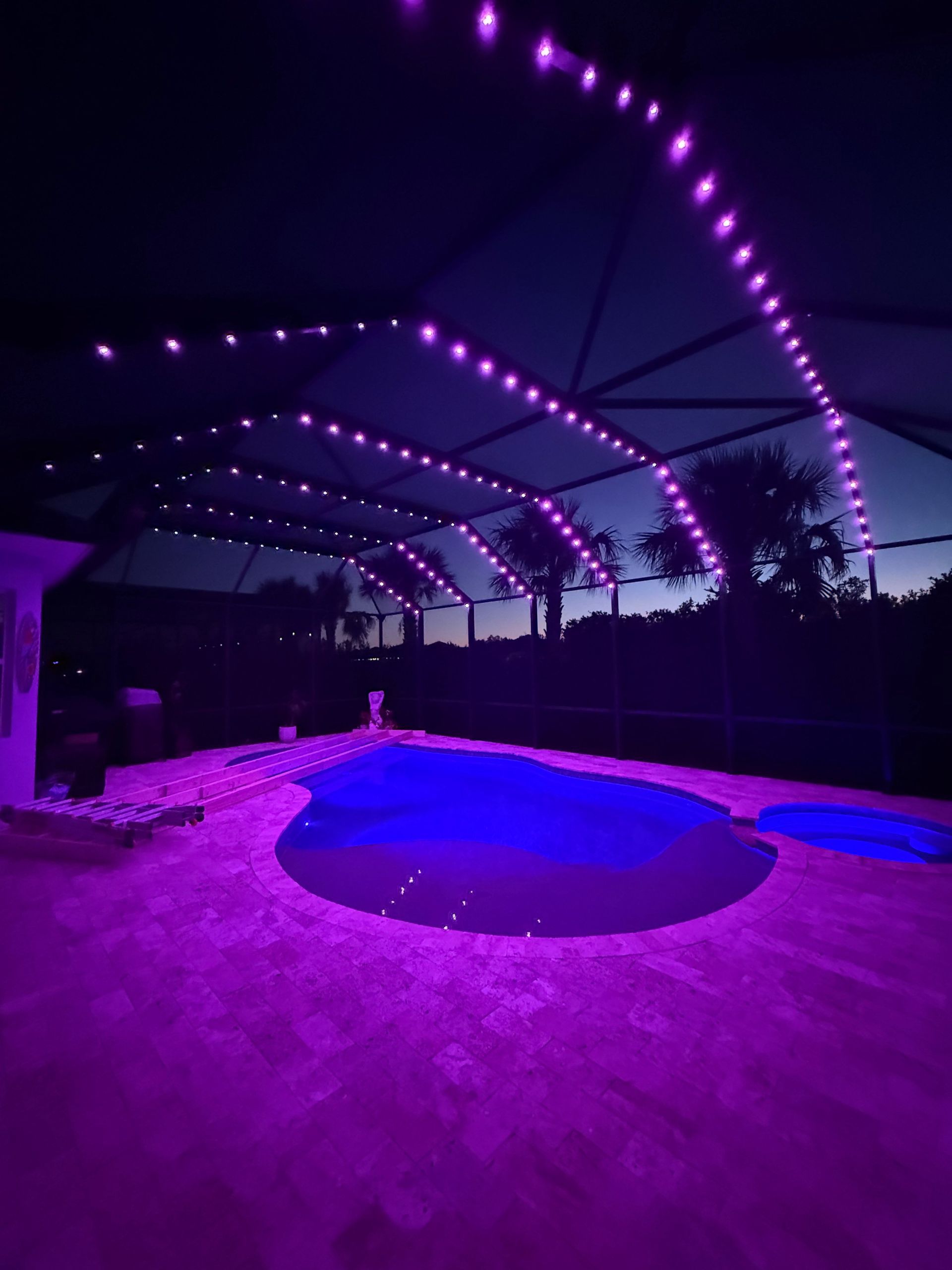Purple string lights illuminate a pool and patio area at dusk.