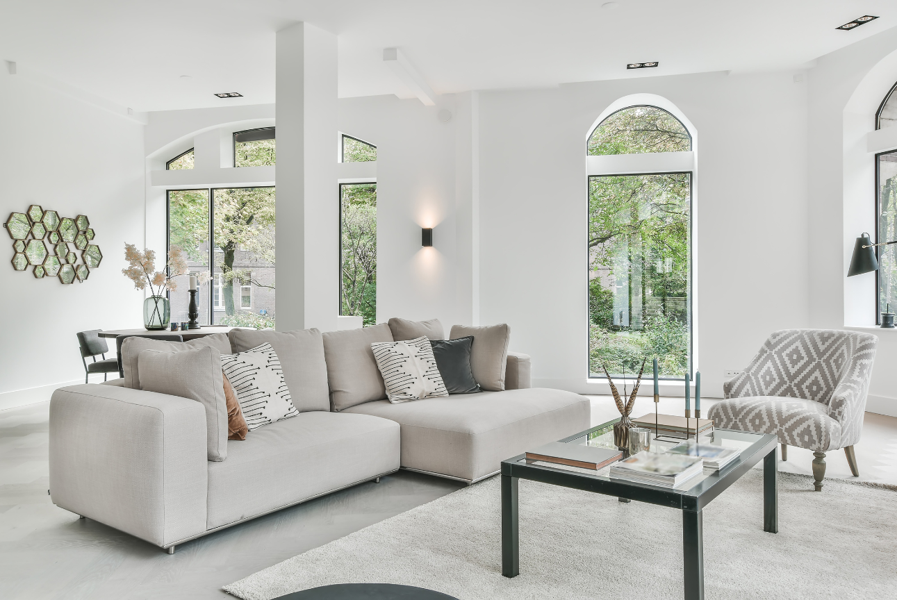 Bright living room with a beige sectional sofa, glass coffee table, and large arched windows overlooking greenery.