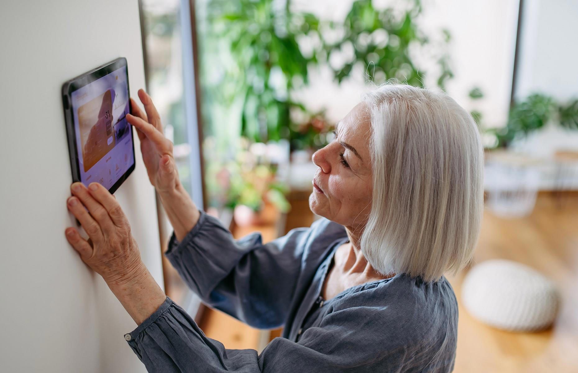 Woman touching a tablet mounted on a white wall; background shows a window with plants.