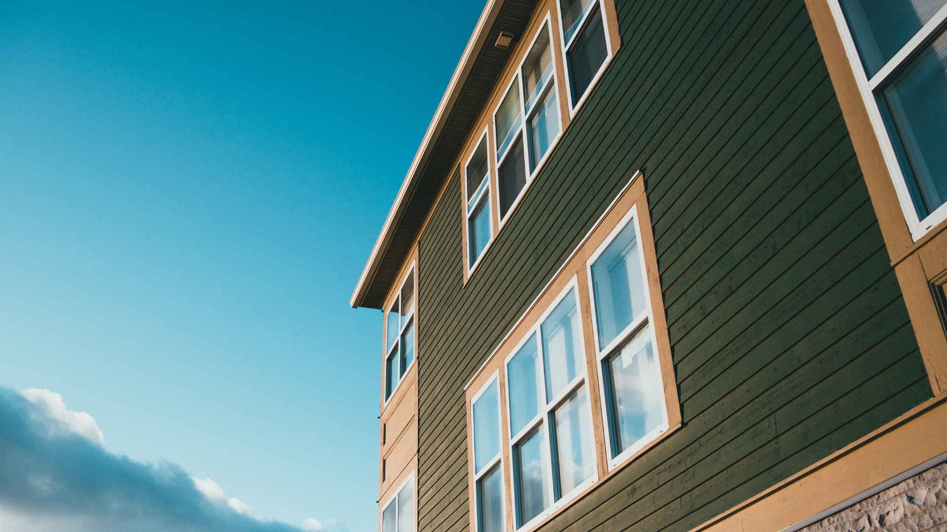 Green and tan building with white-framed windows against a blue sky with clouds.
