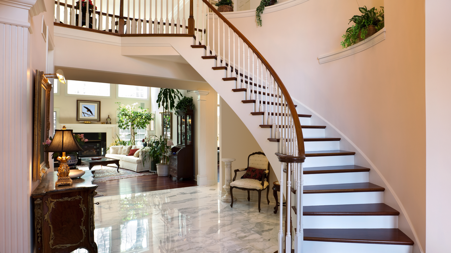 Elegant foyer with curved staircase, marble floor, and view of a living room with fireplace.