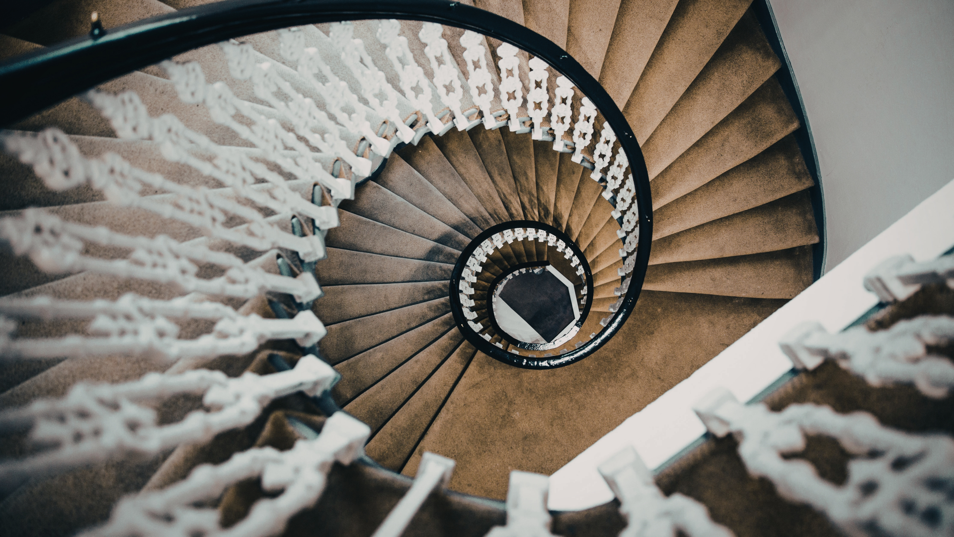 Spiral staircase with brown carpet and white balustrade, viewed from above.