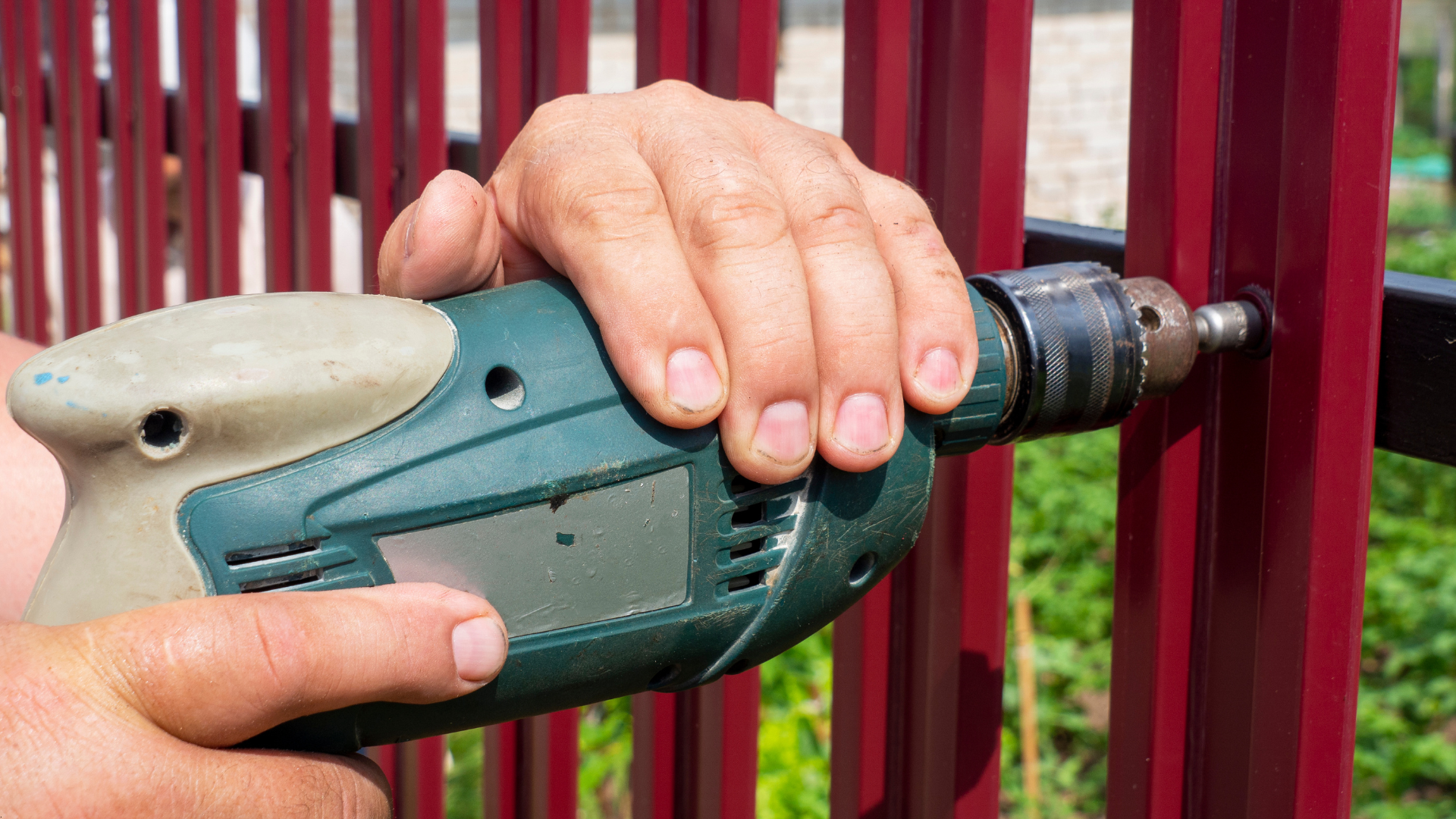 A person using a power drill to fasten something to a burgundy metal fence.