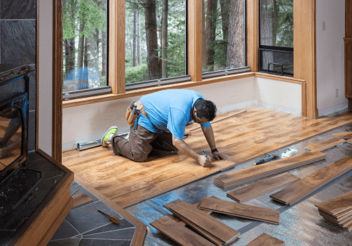 Man installing hardwood flooring in a room with large windows overlooking a forest.