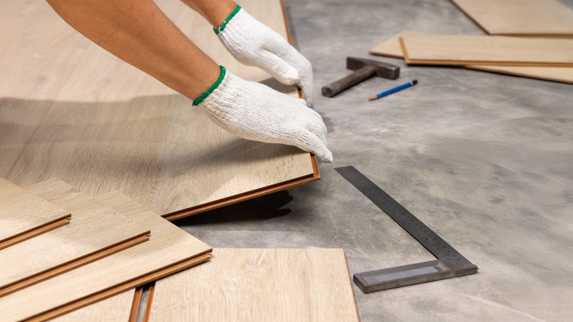 Hands in work gloves installing wooden floorboards, tools nearby.