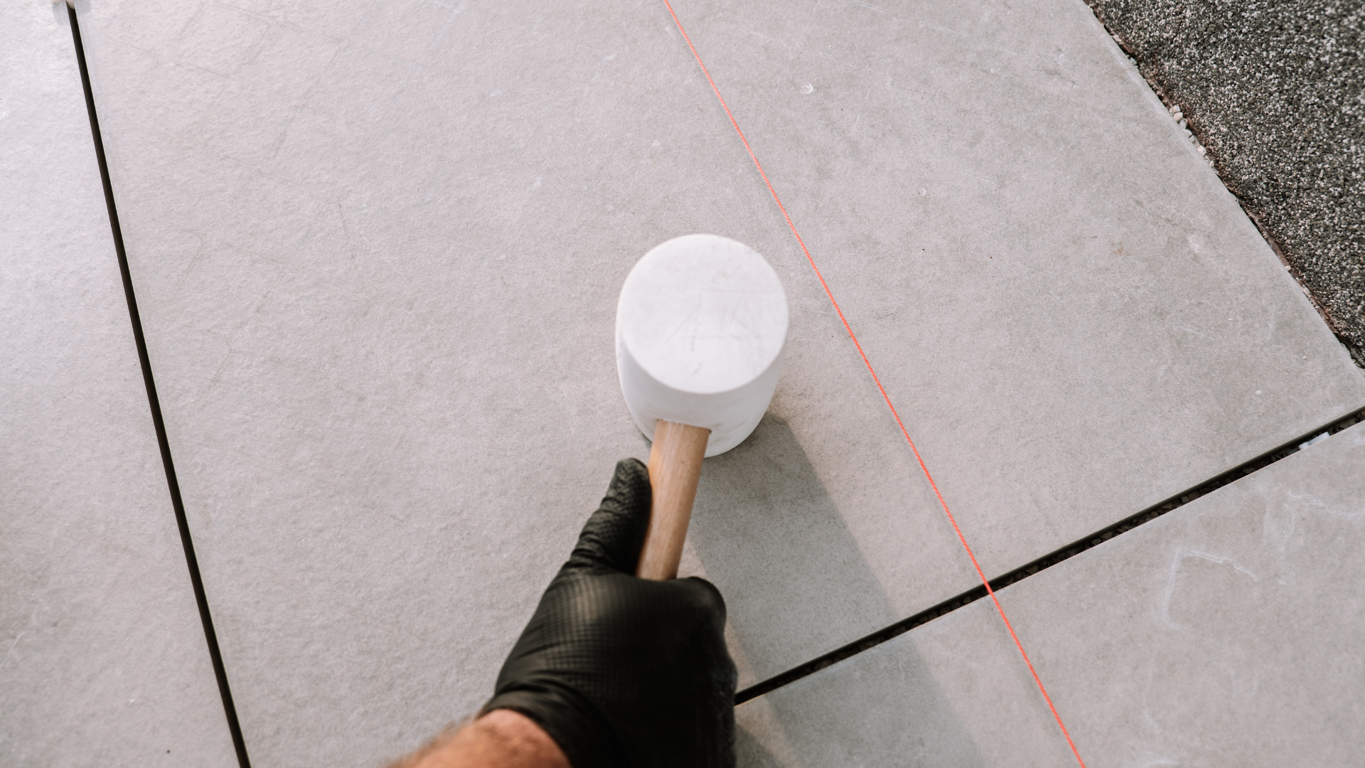 Person in black gloves using a mallet to level a gray floor tile, next to black grout lines.