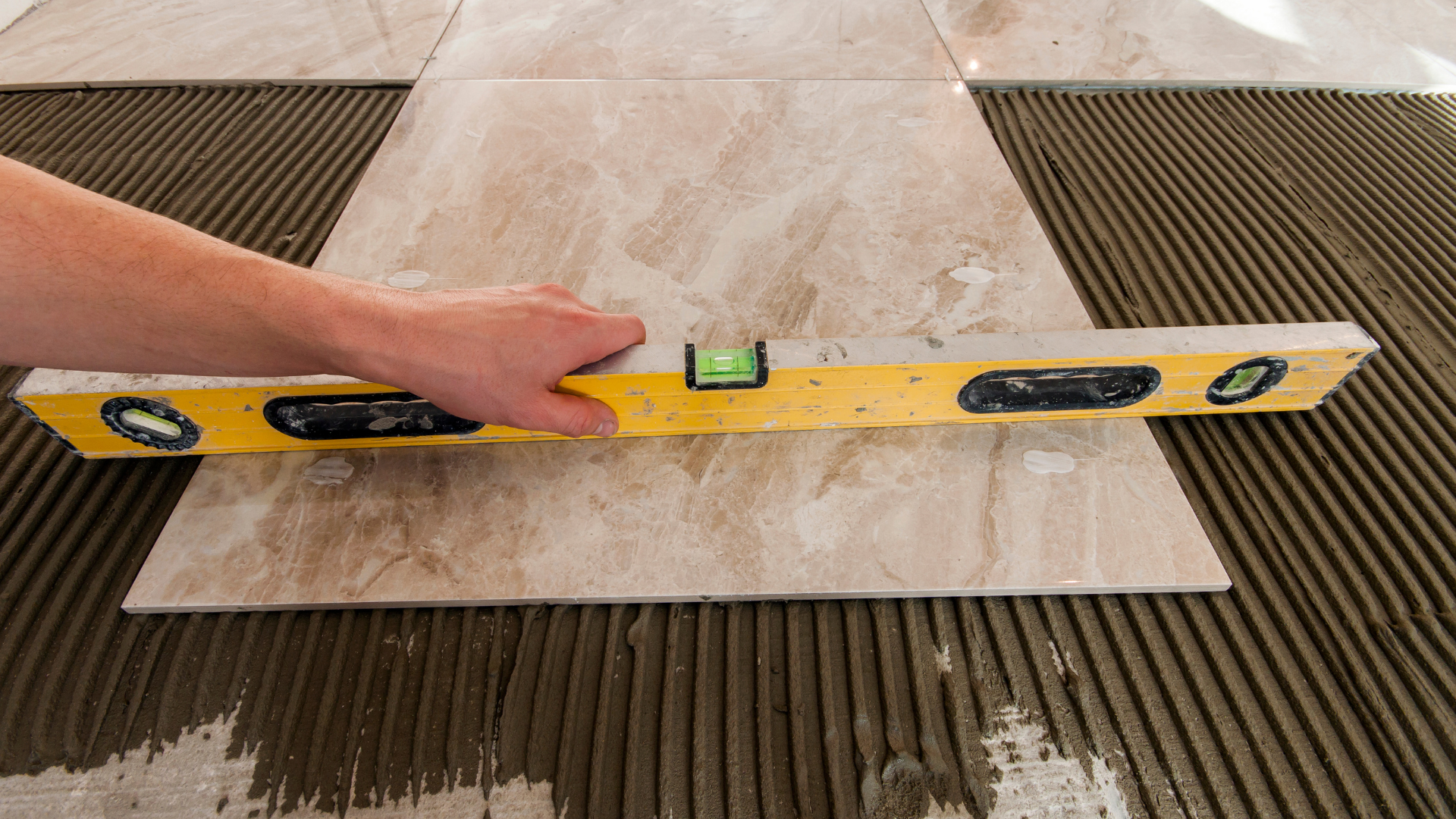 Hand holding a yellow level to check the alignment of a beige floor tile on wet adhesive.