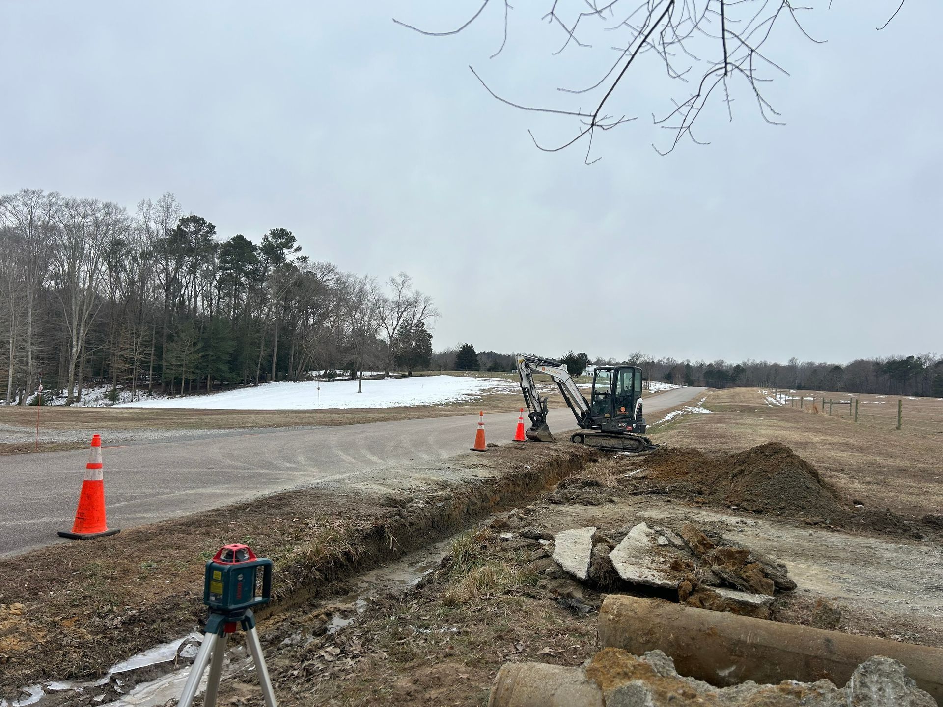 A construction excavator digs a trench along a gravel road on a winter day, with traffic cones and a laser level in view.