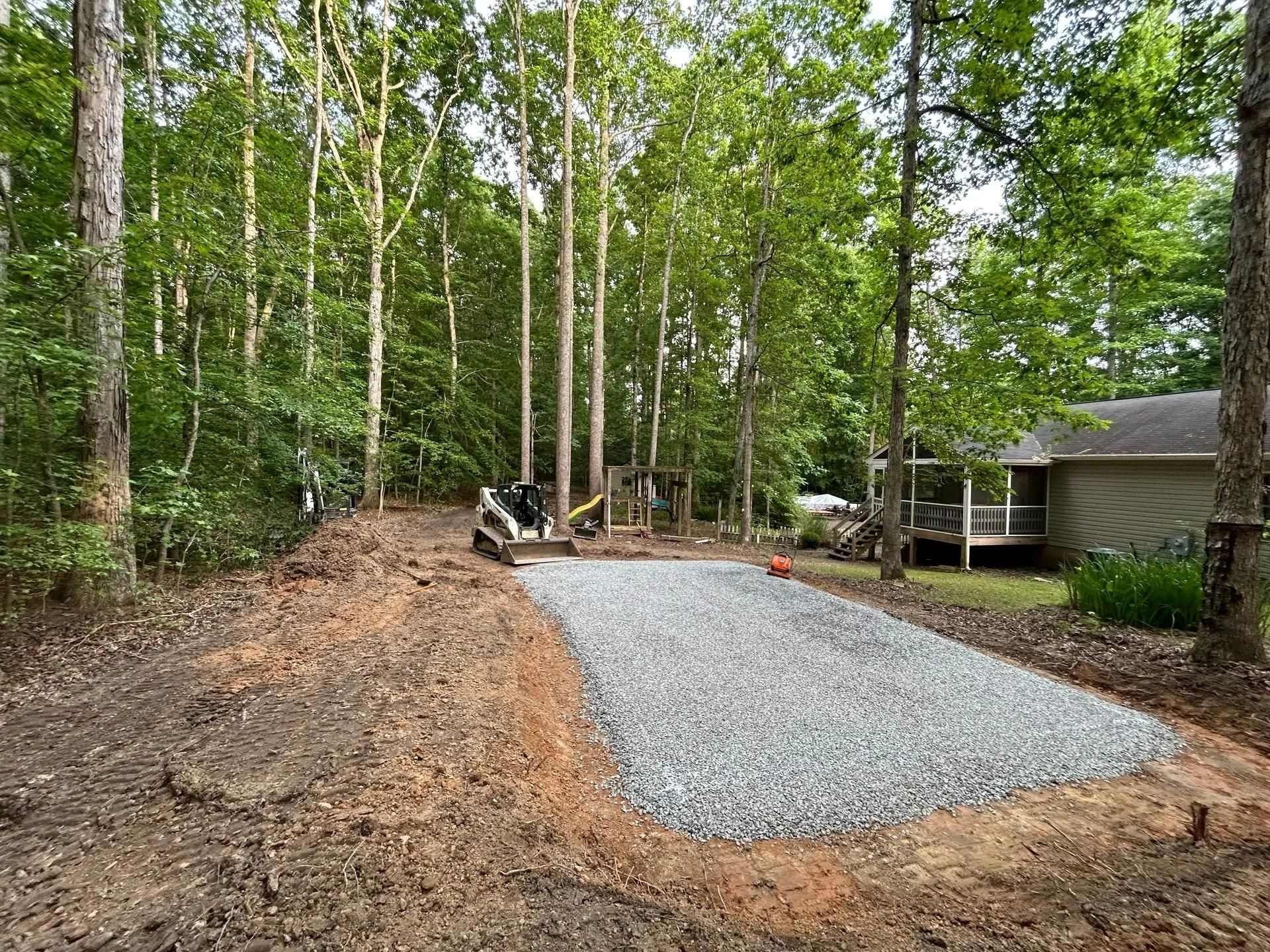 A skid steer loader sits next to a newly laid rectangular gravel driveway in a wooded backyard near a house.