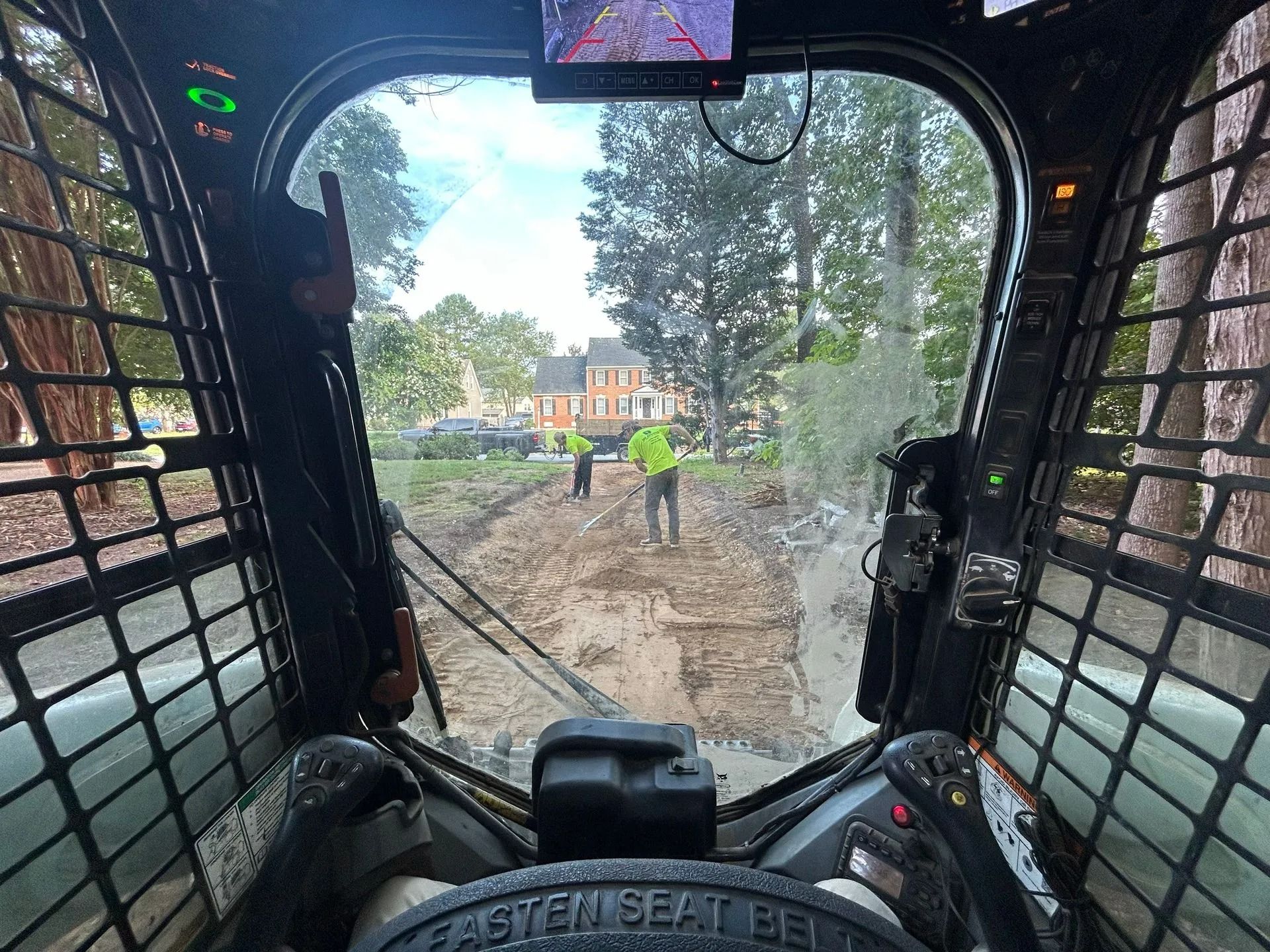 View from inside a skid steer loader as it faces two workers in bright safety vests working on a muddy construction site.