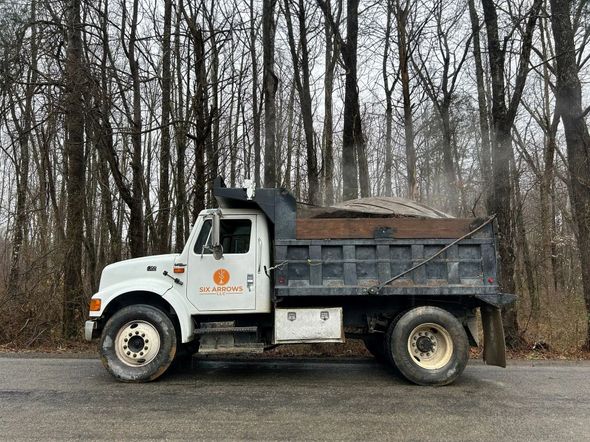 White dump truck with an orange logo parked on a road against a backdrop of bare trees.