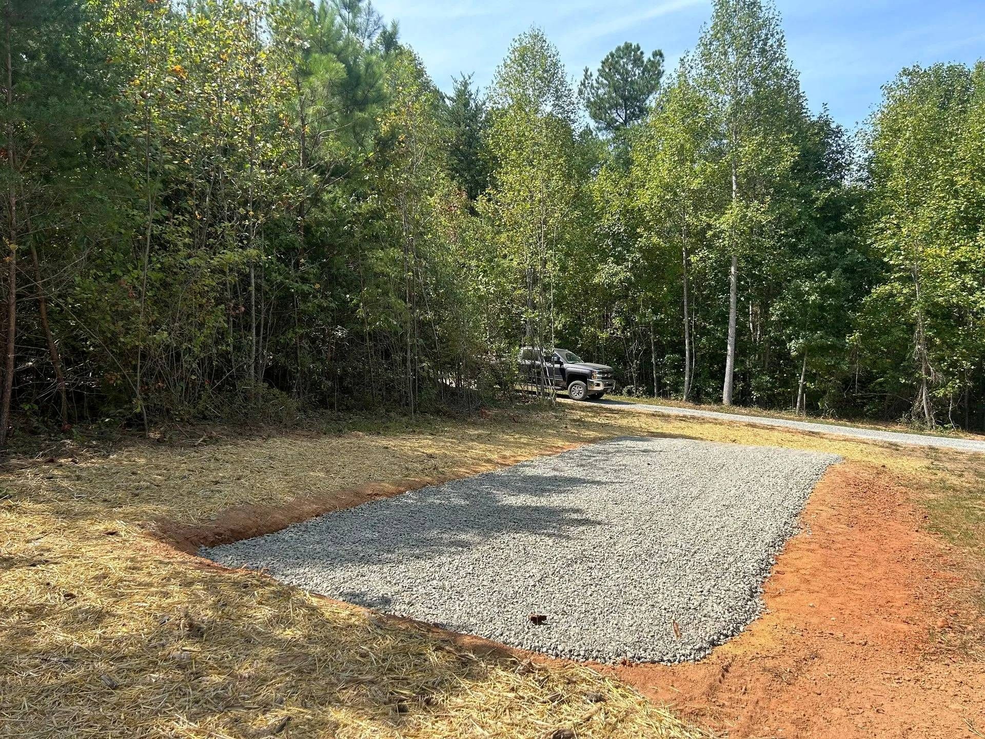 A rectangular gravel pad sits in a cleared, grassy area bordered by a dense forest, with a truck parked in the background.
