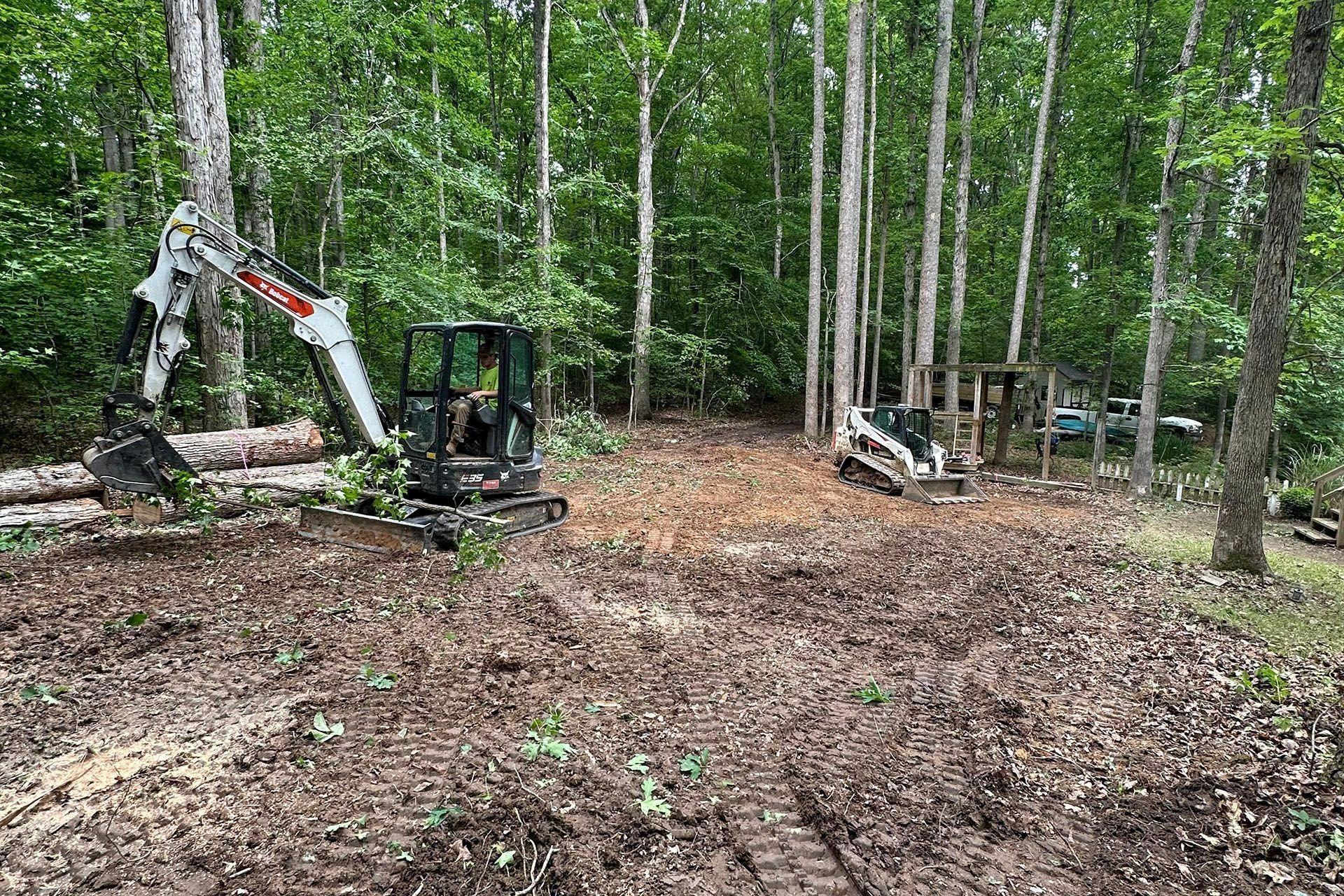 A Bobcat excavator and a skid-steer loader working in a wooded area to clear ground and move logs.