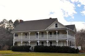 Large white two-story house with wraparound porches and dark roof on a grassy hillside