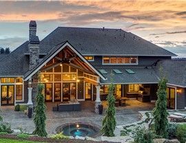 Large gray house with a stone patio, pool, and trees, viewed at dusk.