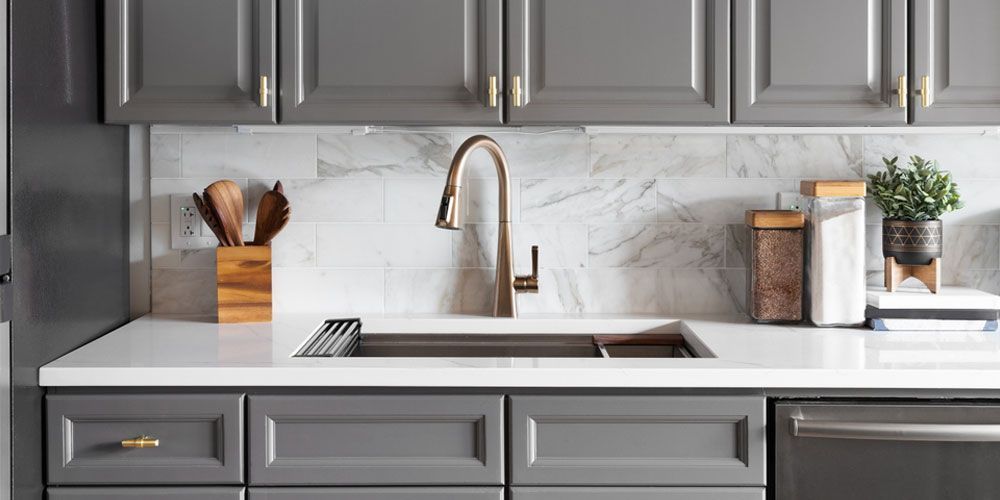 Kitchen with gray cabinets, white countertop, marble backsplash, and bronze faucet.
