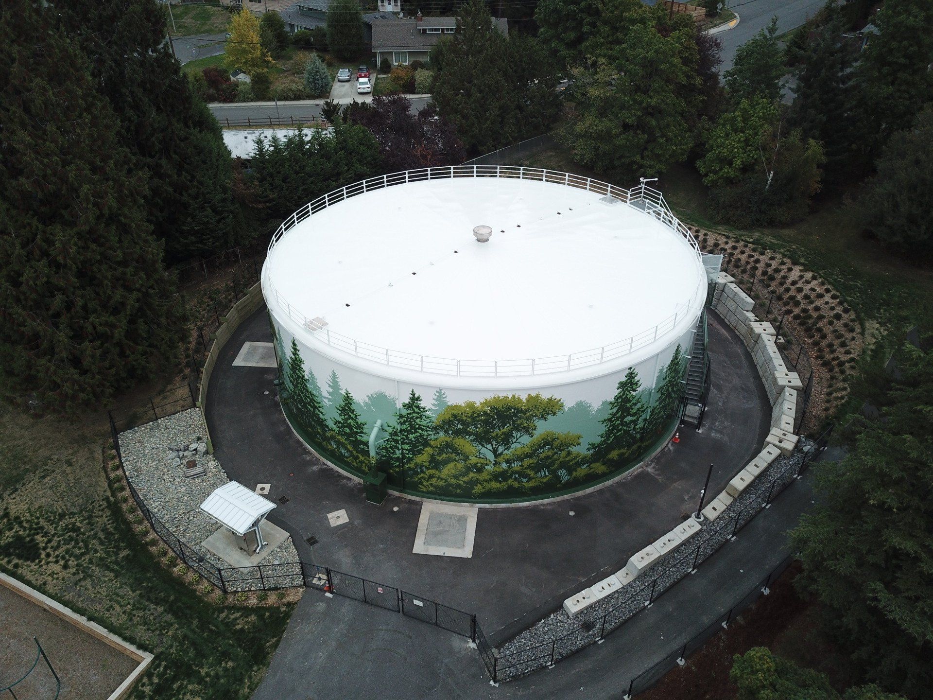 Overhead view of a white water tank with a painted forest scene; circular, with a black fence and stone retaining wall.