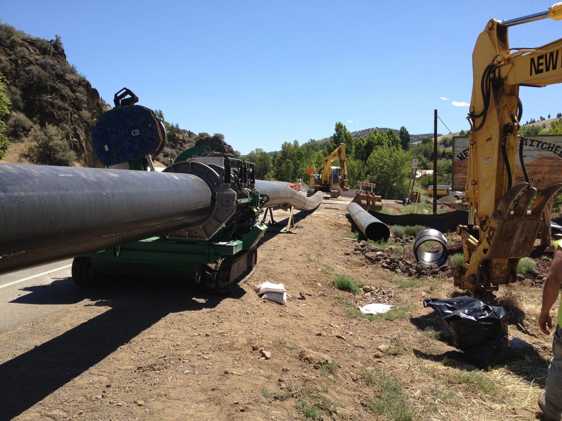 Construction site: large pipe being joined, road, excavator, sunny day.