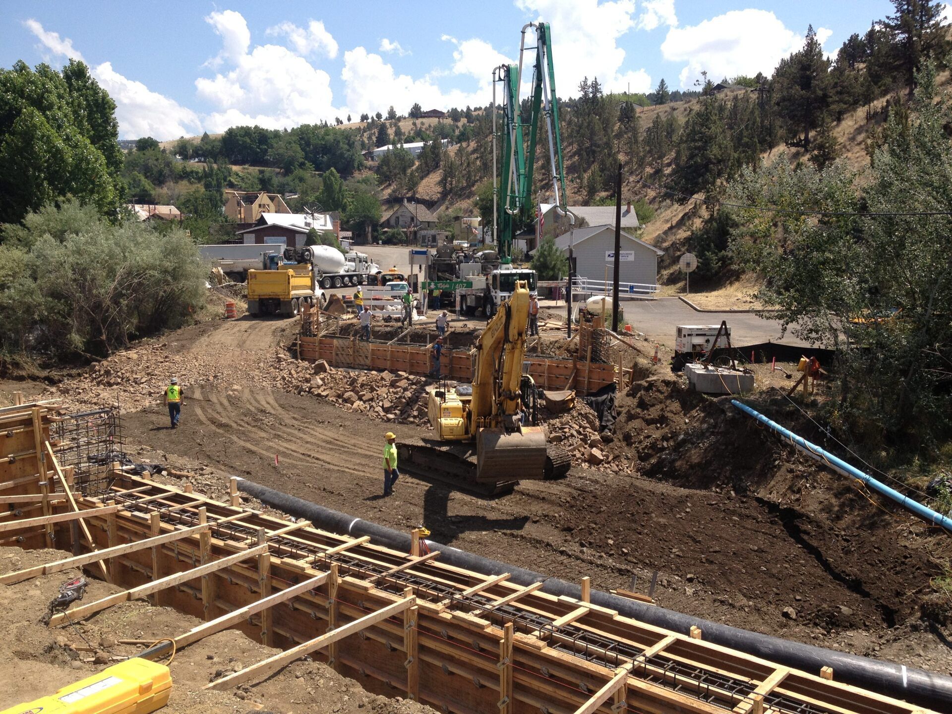 Construction site: Excavator, concrete pump, workers, wooden forms, dirt, and machinery.