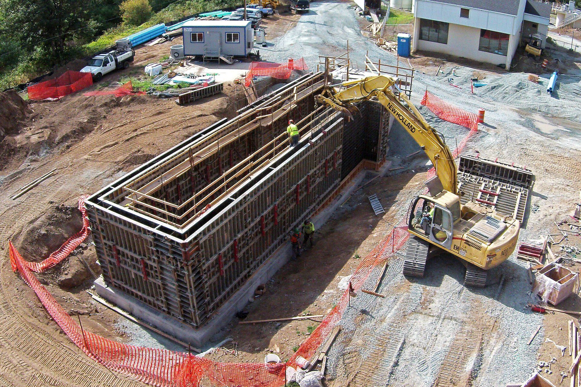 Construction site: Excavator near a rectangular concrete form. Orange safety fencing surrounds the work area.
