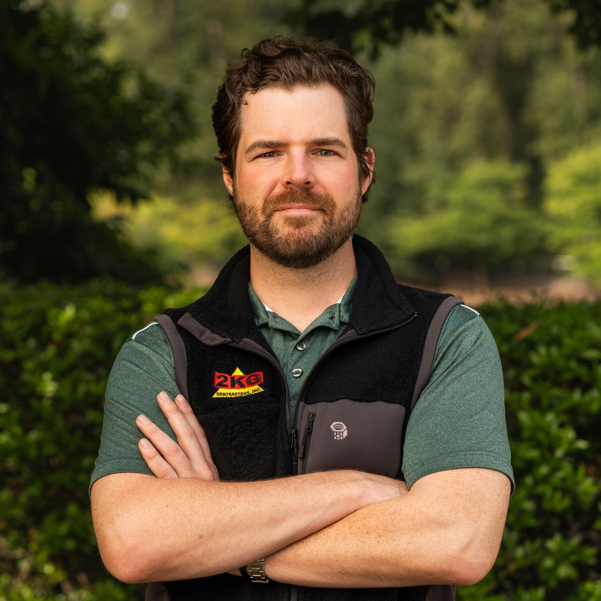 Man with beard and crossed arms, wearing a green shirt and black vest, standing outdoors.
