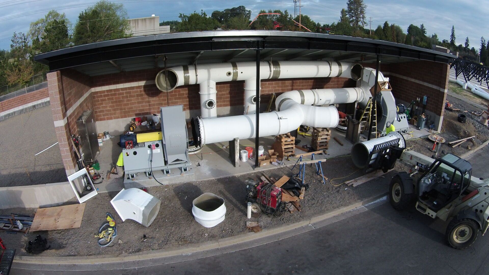 HVAC system being installed outdoors; large white pipes and machinery, on pallets, brick wall.