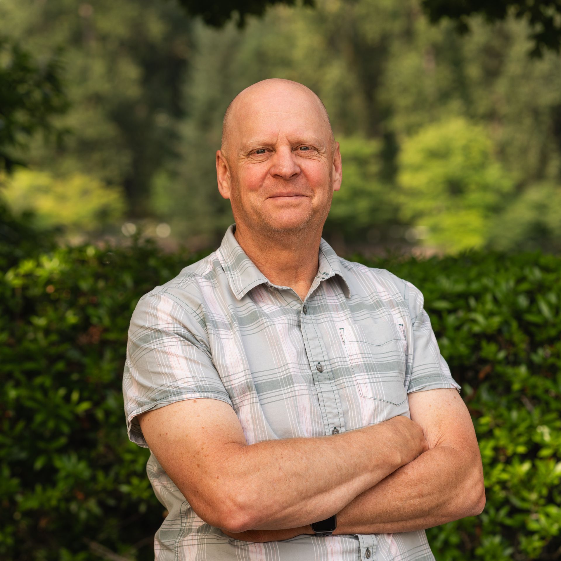 Bald man with crossed arms, smiles at the camera, wearing a plaid shirt. Outdoors with green foliage.