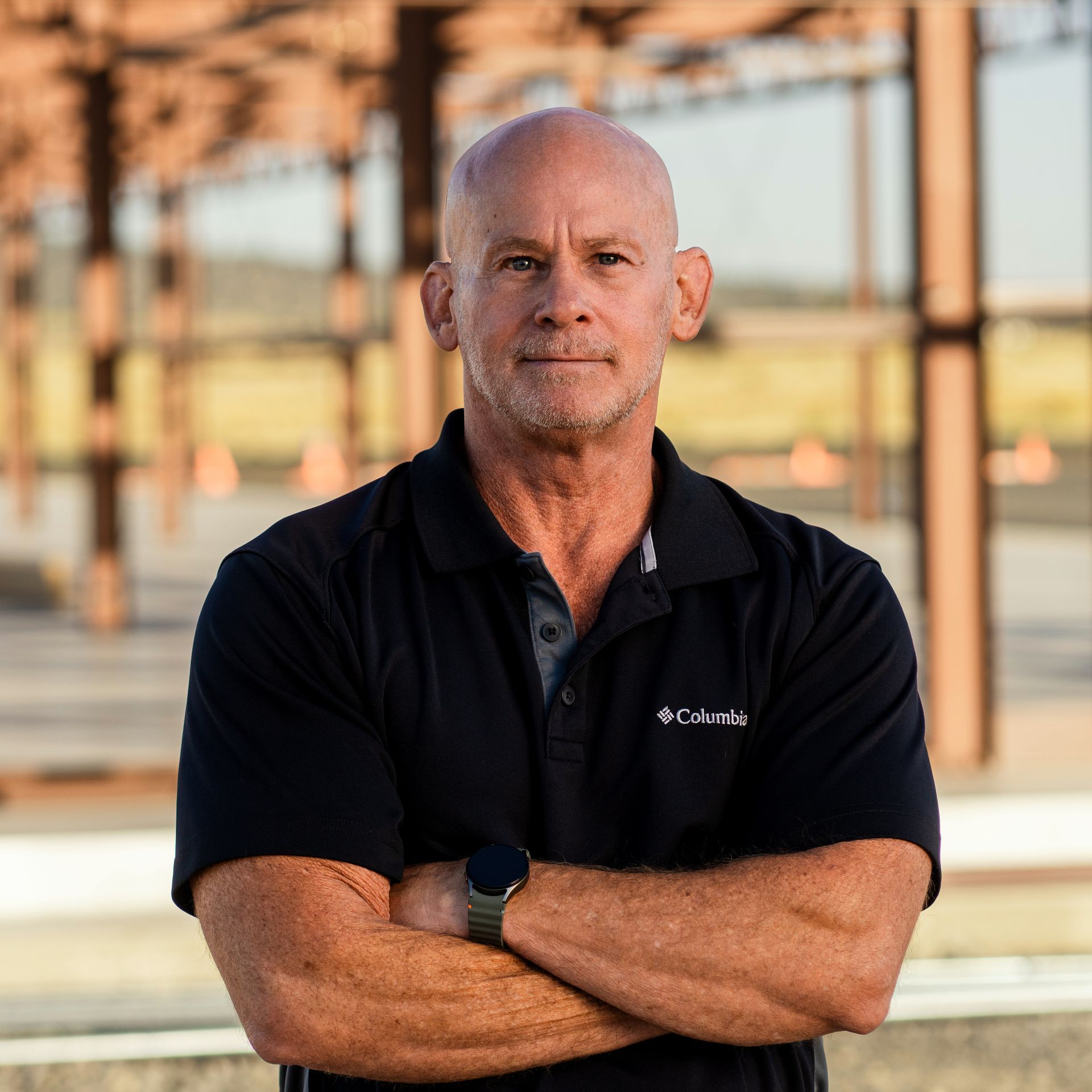 Bald man with arms crossed, wearing a black shirt with a Columbia logo, standing outside.