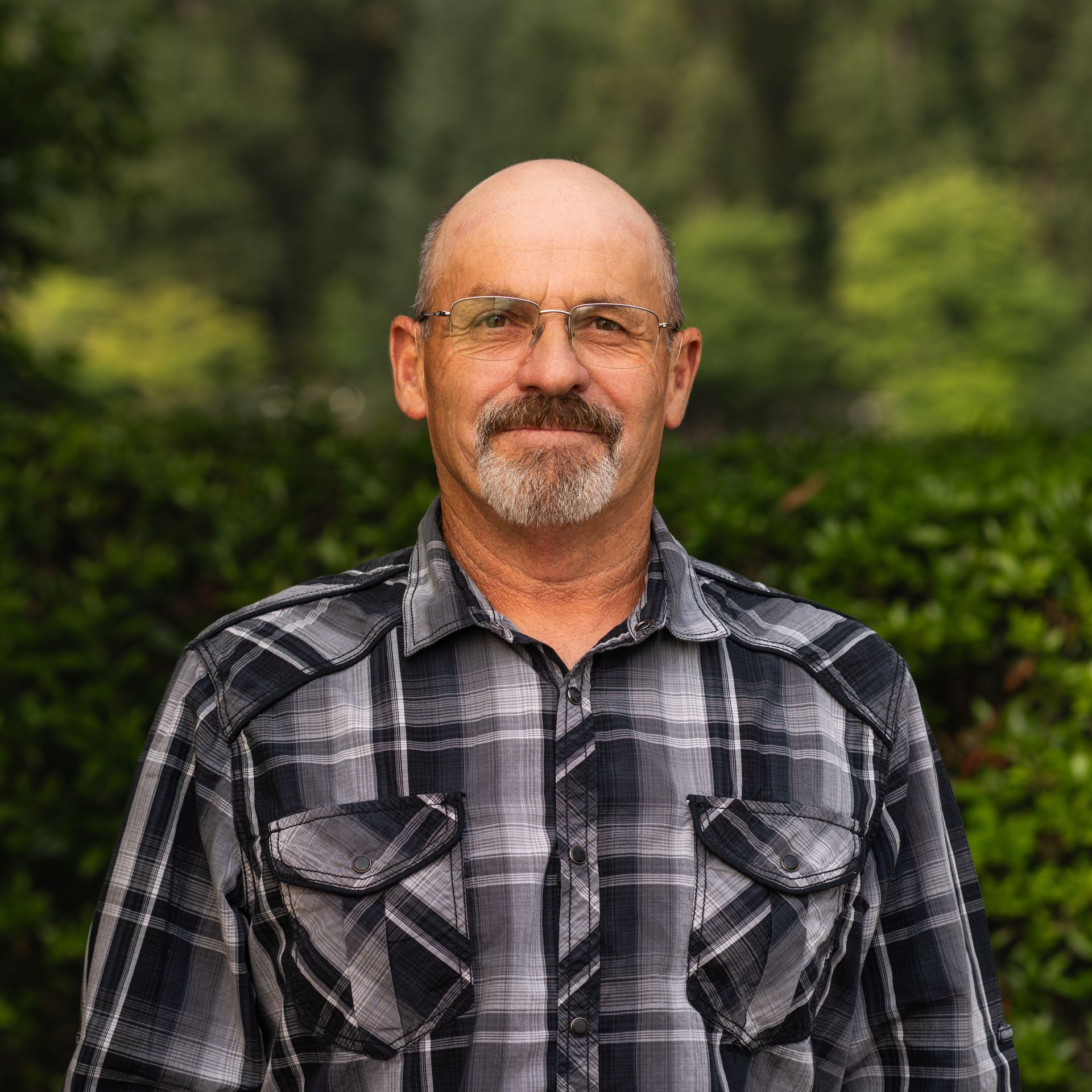 Bald man with glasses, goatee, and plaid shirt against a blurred green background.